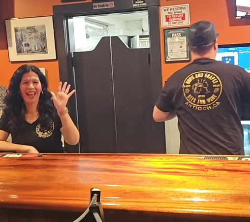 A woman smiling and waving behind a wooden counter, with a man facing away working on a computer. The scene appears to be inside a business or office.