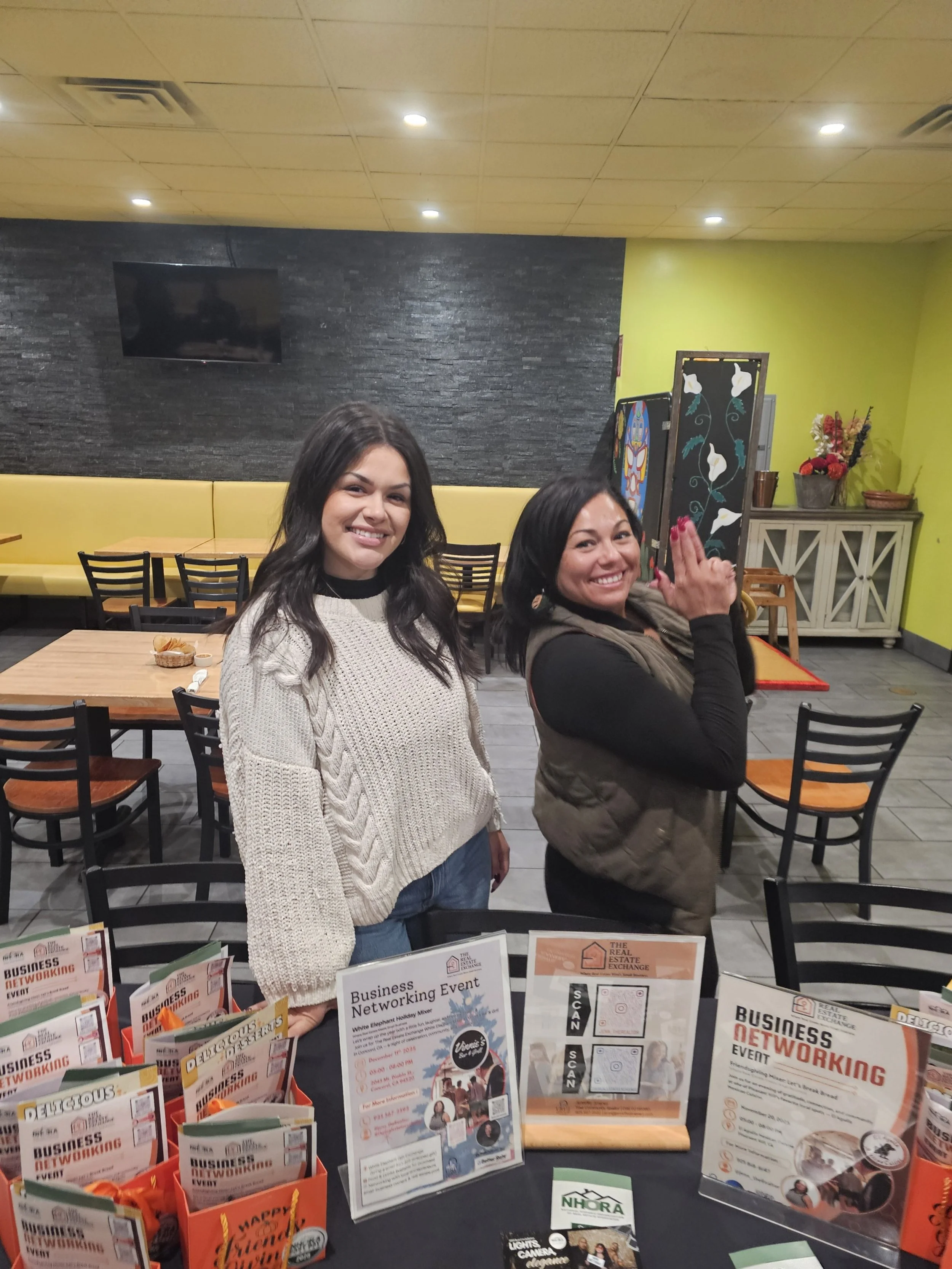 Two women standing behind a table at a business networking event in a restaurant or cafe setting. The table has informational flyers and promotional materials related to the event.