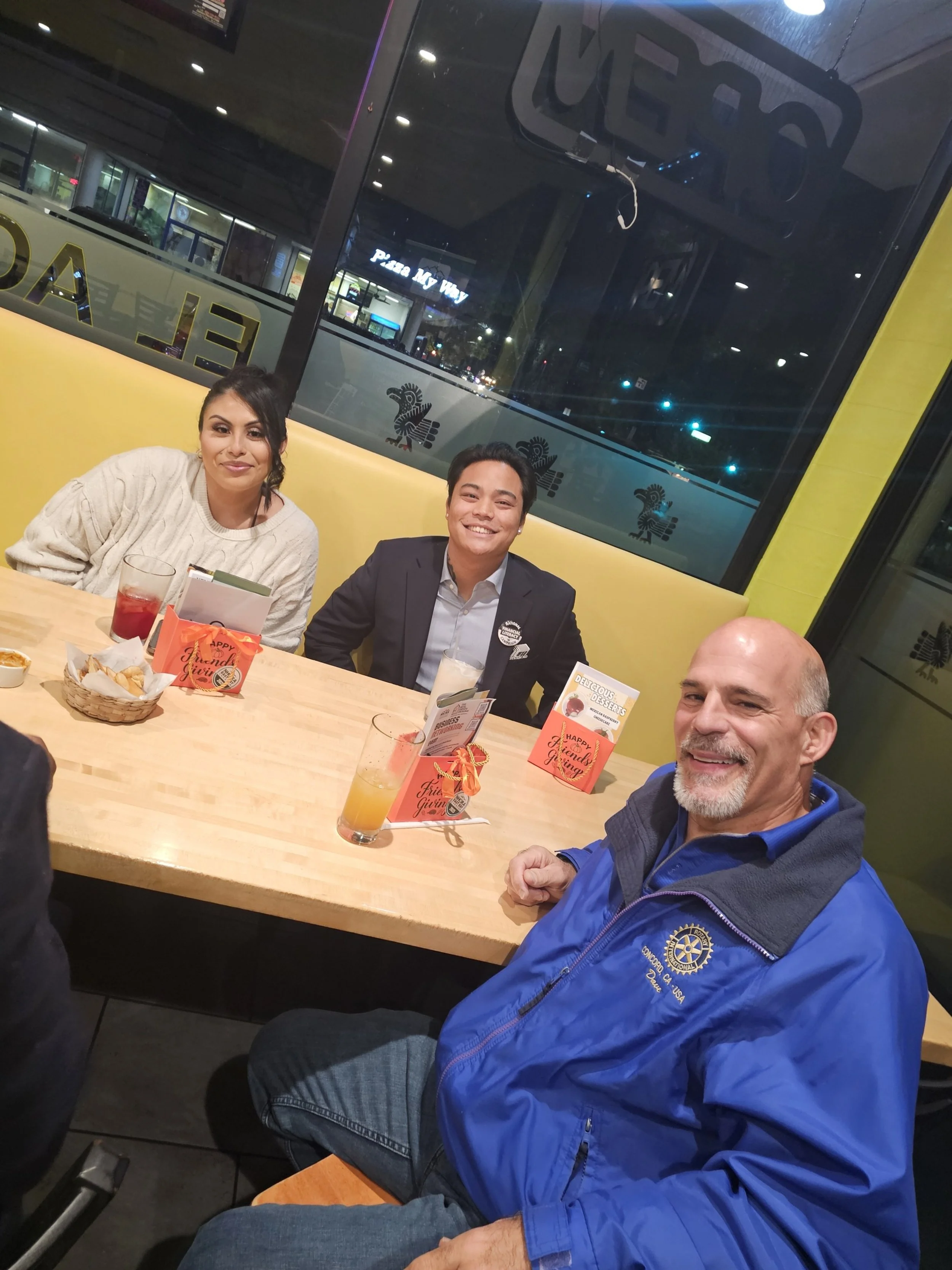 Three people are sitting at a table inside a restaurant, smiling at the camera. Two women and one man. The woman on the left has dark hair and is wearing a cream sweater. The man on the right is bald with a beard and is wearing a blue jacket. The wom