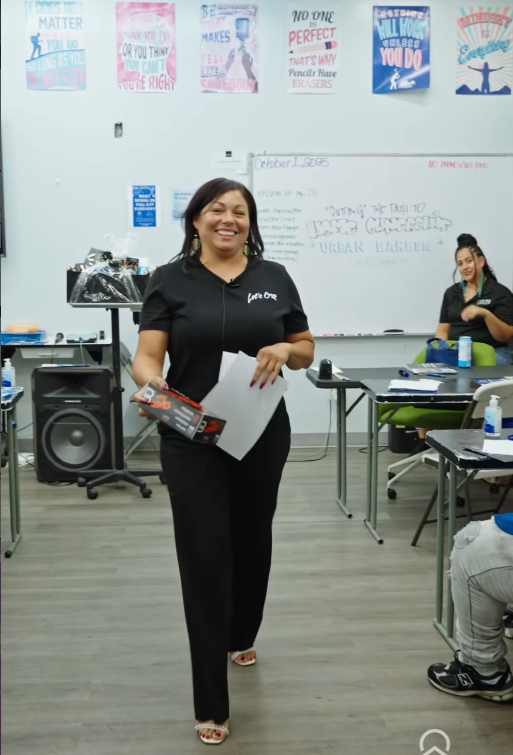 A woman smiling and holding papers and a boxed item in a classroom or meeting room with desks, posters, and another person seated in the background.