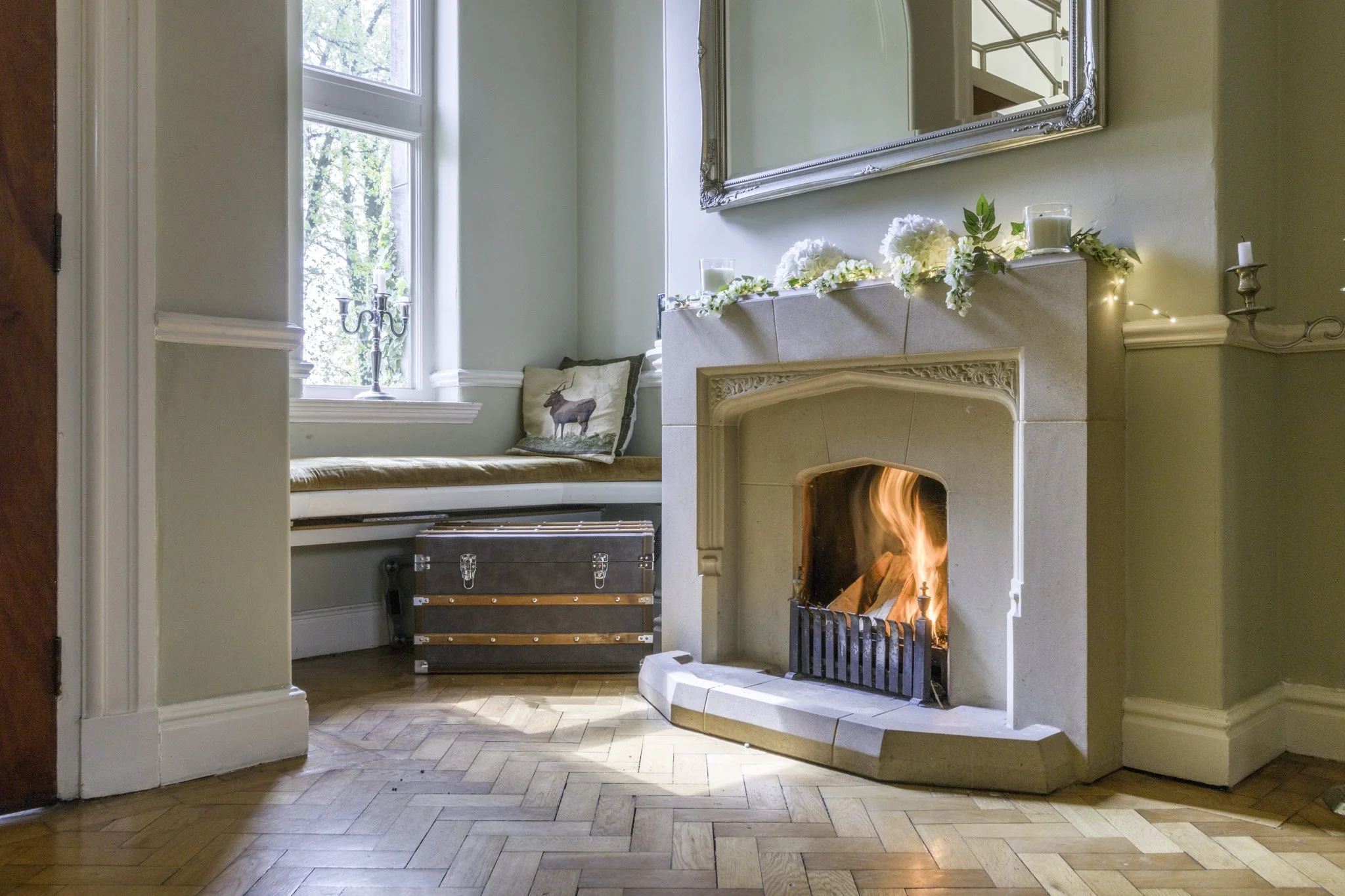 Cozy living room with a roaring fireplace, decorated with white flowers and candles on the mantel, a window with a view of trees, and a cushioned window seat with a pillow with a moose design. There is a vintage trunk on the hardwood floor.