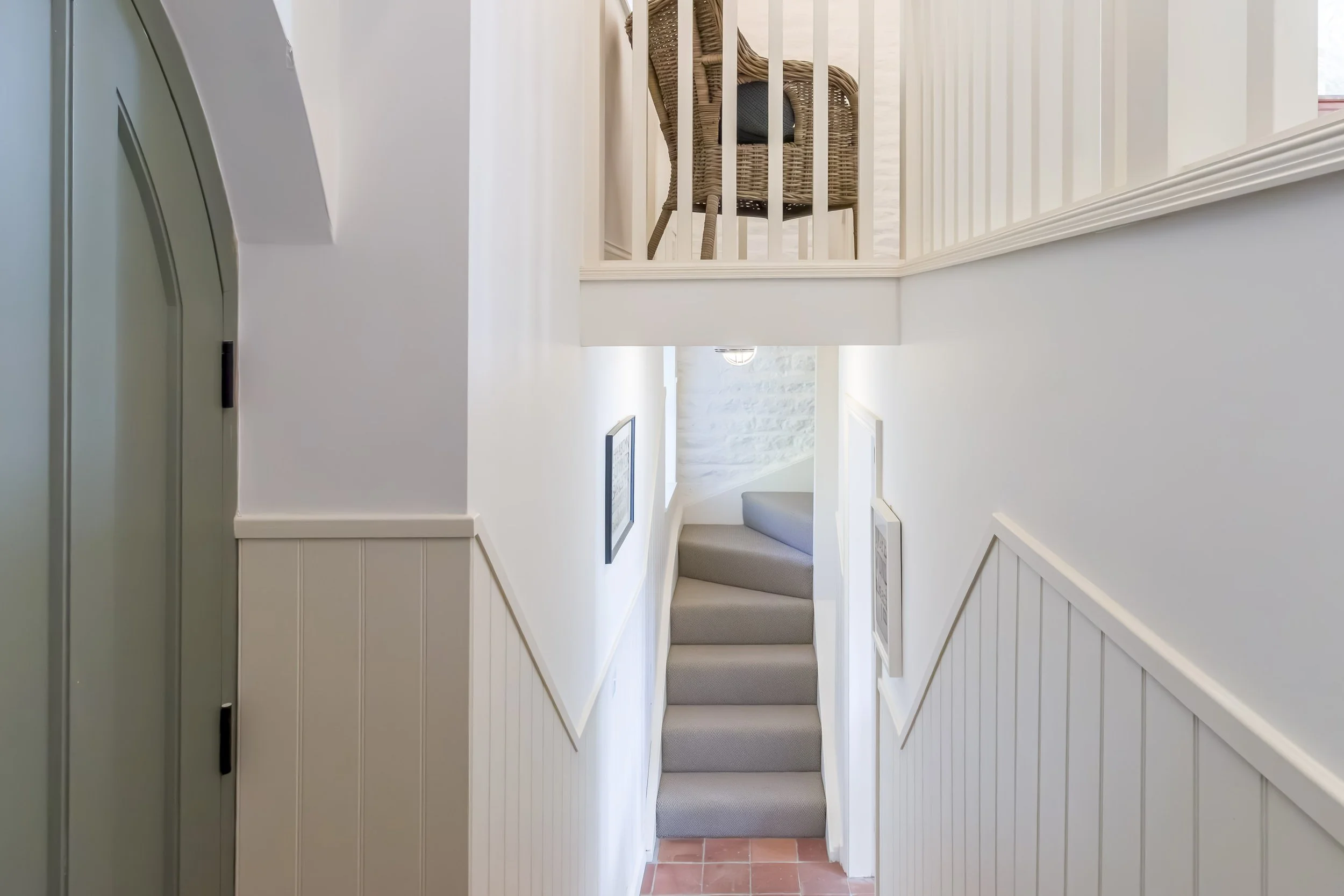 Interior view of a narrow staircase with carpeted steps, white walls, and framed pictures, leading up to a small landing with a wicker chair placed against a white railing.
