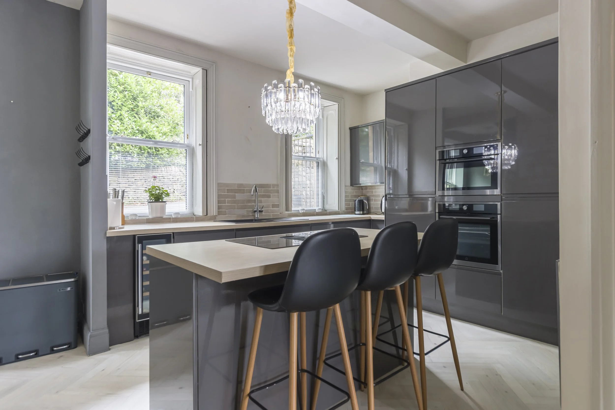 Modern kitchen with gray cabinets, a central island with a beige countertop, three black barstools, two windows, a chandelier, and built-in oven and microwave.