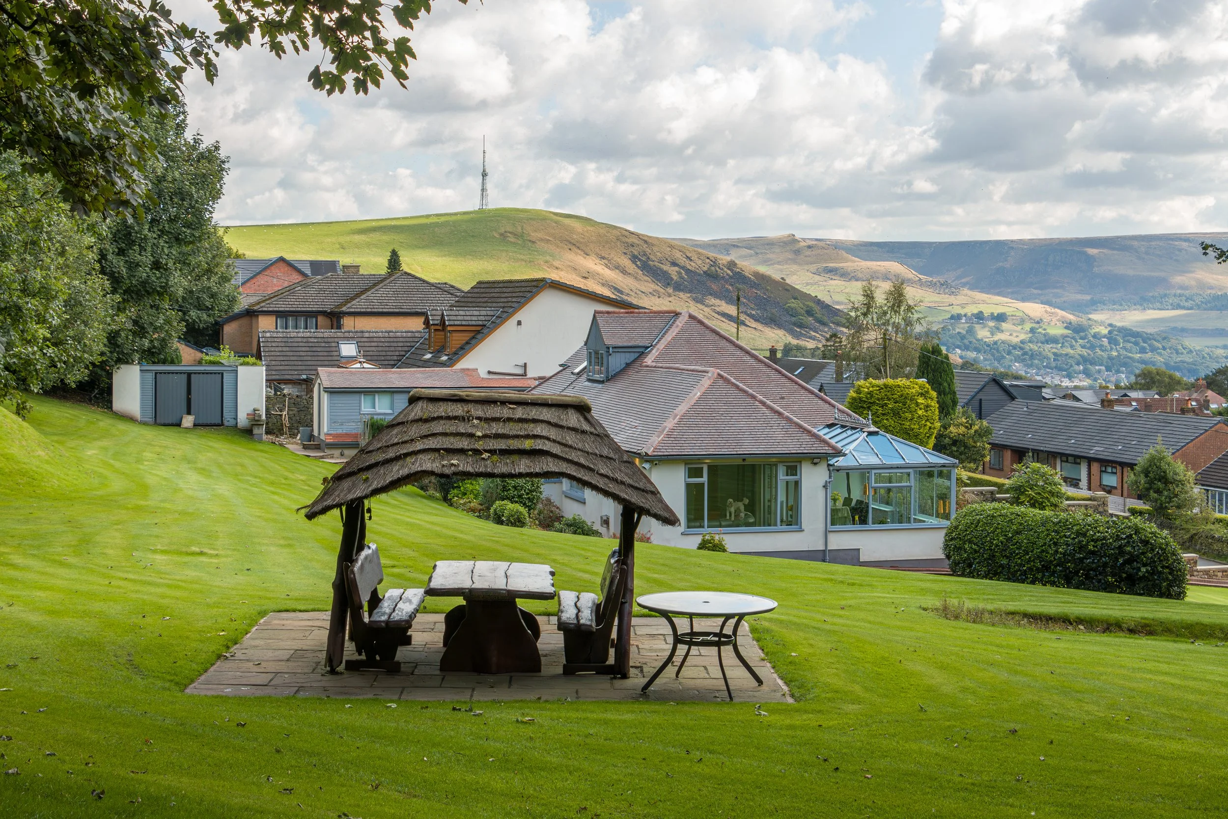 Lawn with wooden gazebo and table in front of houses on a hillside, with rolling hills and cloudy sky in the background.