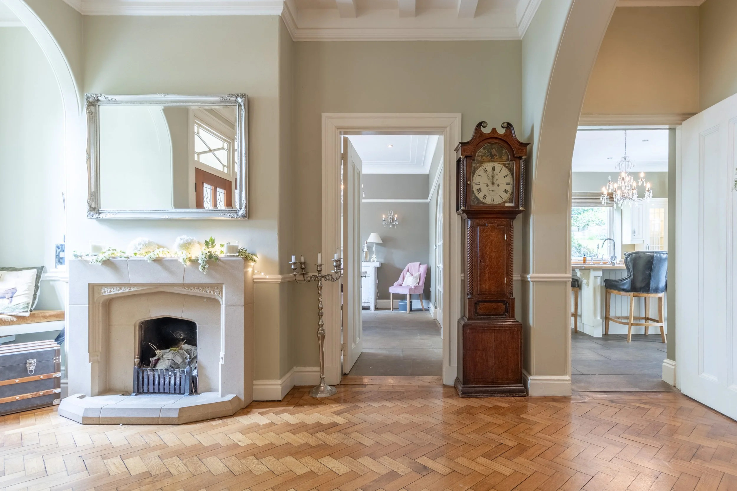 Living room with fireplace, vintage clock, mirror, candelabra, and hardwood floor, leading to kitchen and an adjacent room with pink armchair and chandelier.