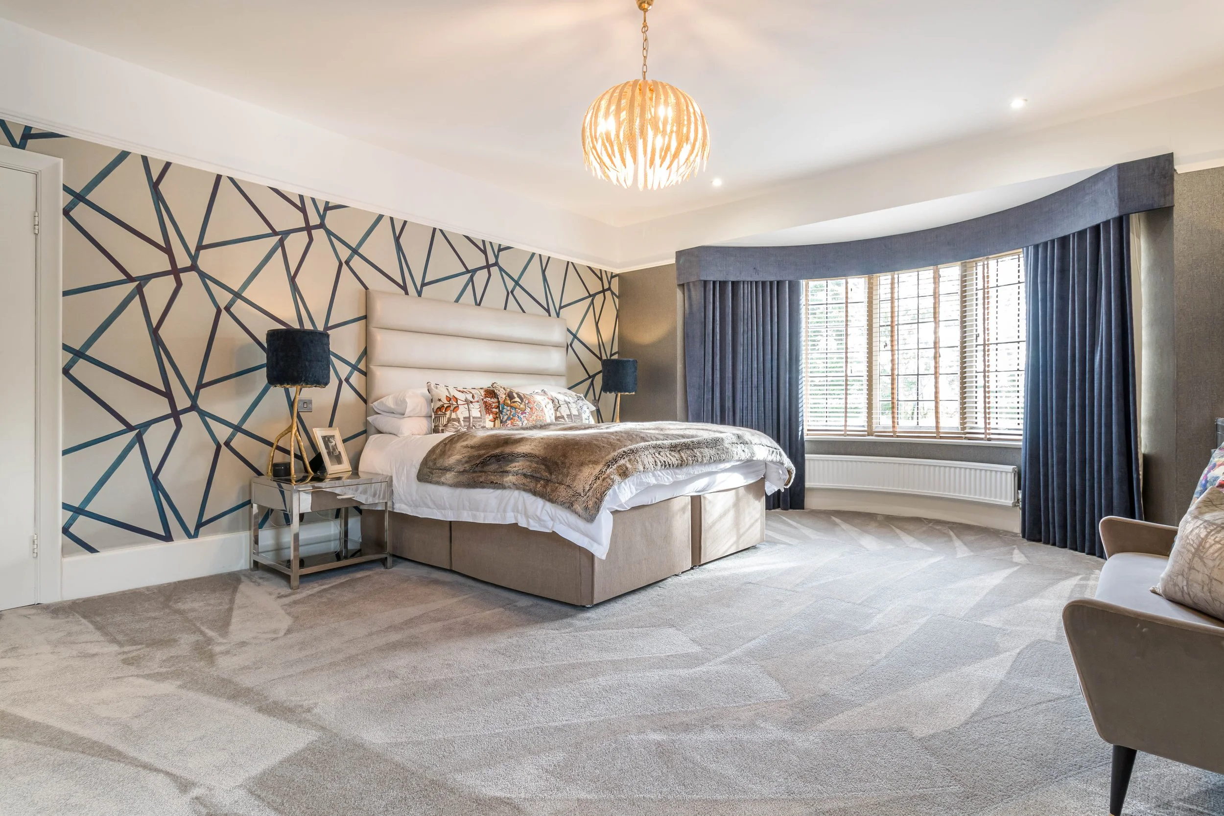 A spacious bedroom with a large bed featuring a tufted headboard, decorative pillows, and a fur throw. There are two black lamps on bedside tables, a geometric-patterned feature wall behind the bed, and a bay window with wooden blinds and navy curtai
