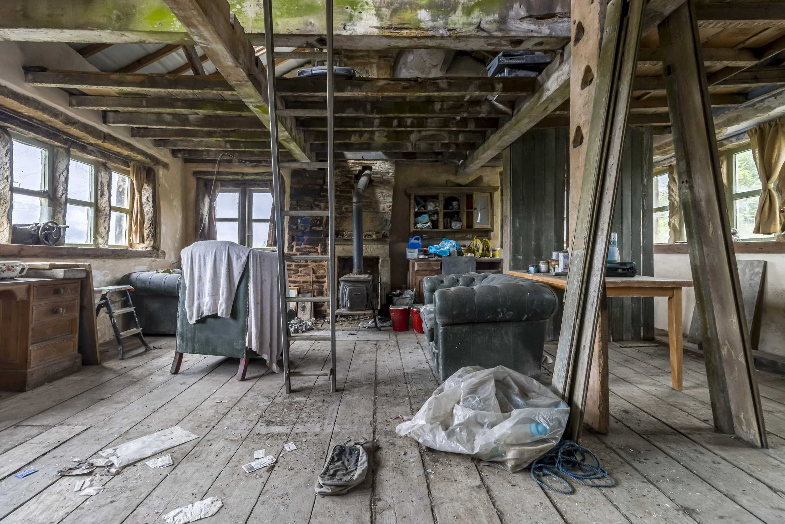 Interior of an old, rustic room with wooden floors, exposed beams, and multiple windows. The room is cluttered with furniture, including a green armchair, a wooden desk, and a cabinet. There are various tools and household items scattered around, and a pile of trash and debris on the floor. A wood stove is in the back corner, and a ladder leans against a support beam.
