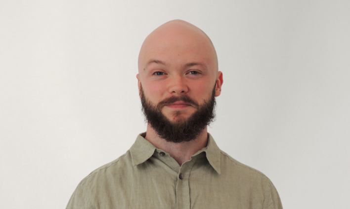 A headshot of owner, Nigel Barnim, bald headed, full beard, and mustache, wearing a beige button-up shirt, standing against a plain white background.