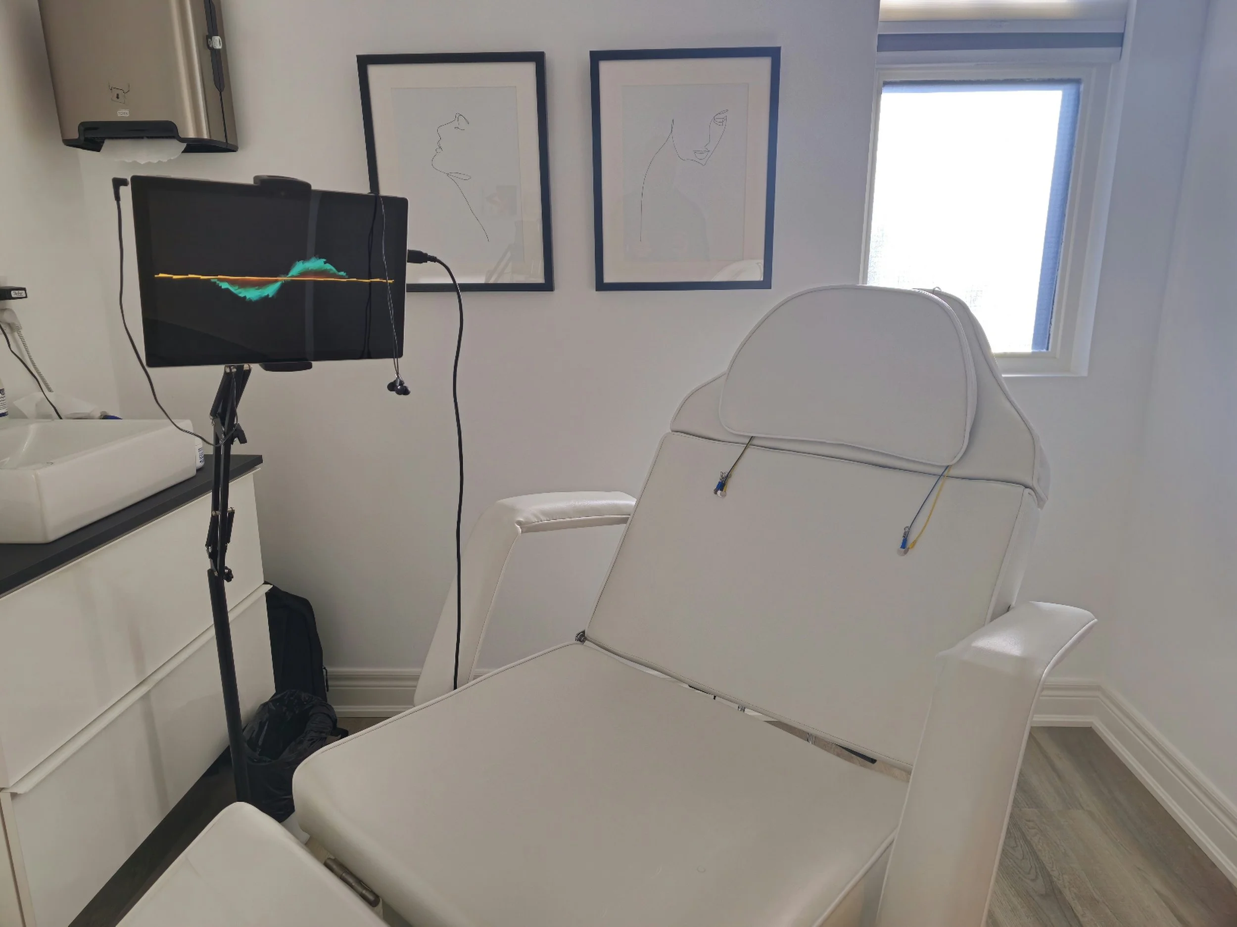 The Neurofeedback treatment room at the Hamilton Clinic, a comfortable recliner chair, a monitor showing an EEG wave, framed line art of faces on the wall, a window, and a small sink.