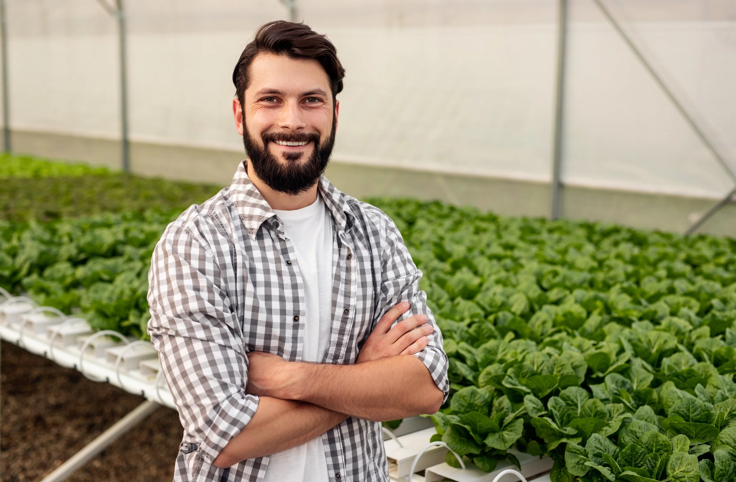 Hombre sonriendo en un invernadero con brazos cruzados, rodeado de plantas verdes.