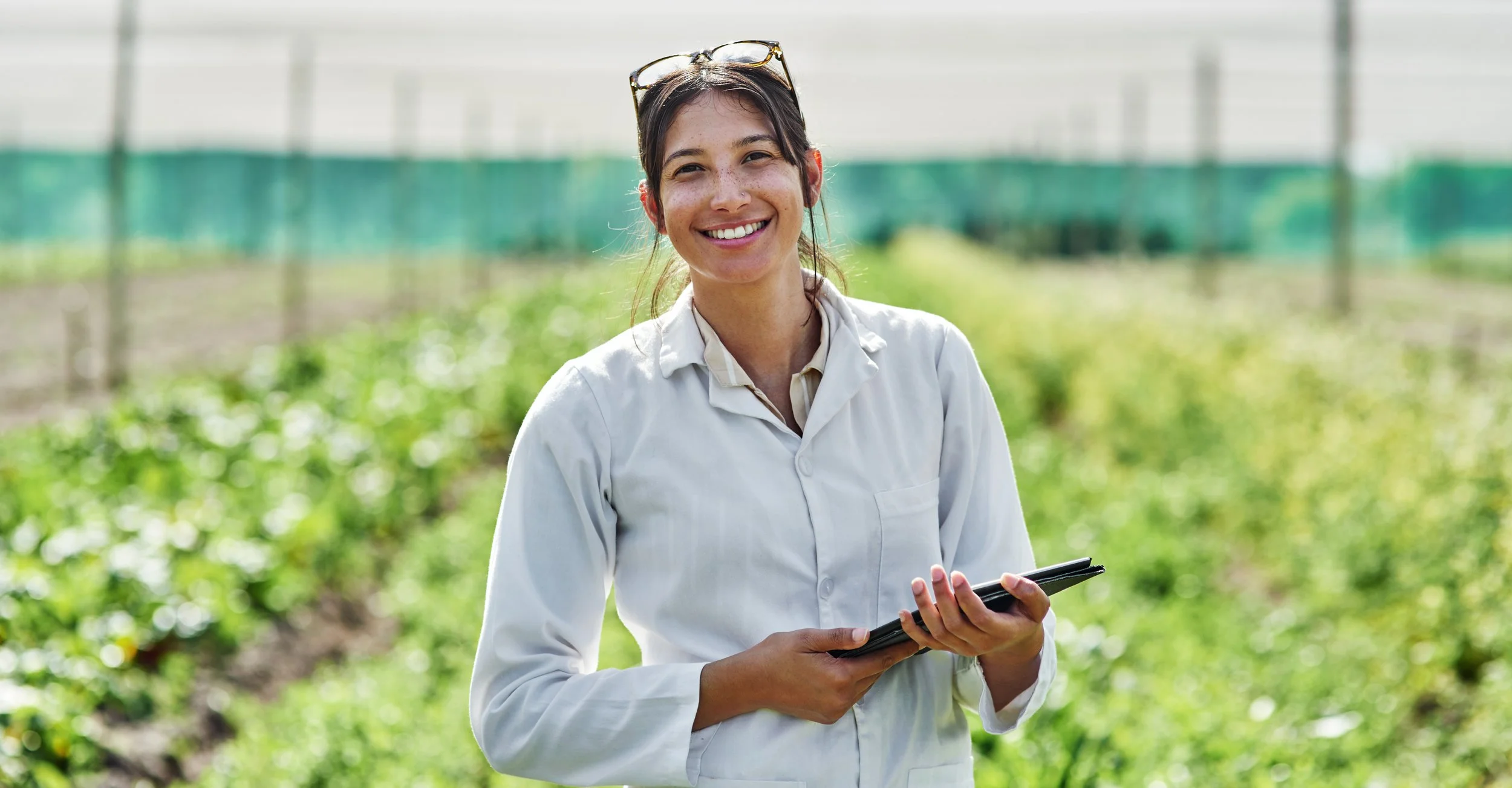 Mujer sonriente en un campo agrícola, vistiendo una bata blanca y sosteniendo una tableta electrónica.