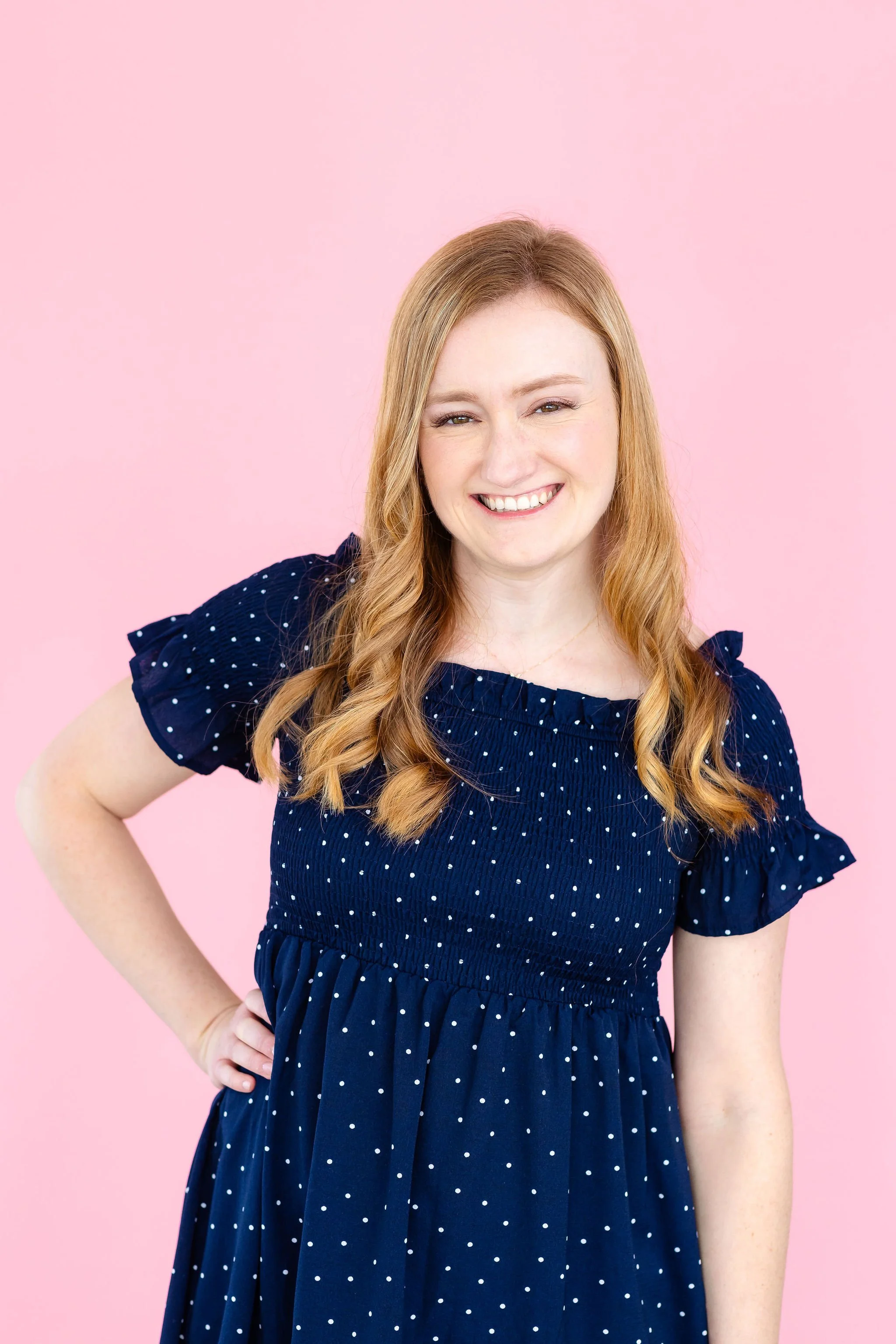 Smiling woman with blond hair wearing a blue polka dot dress against a pink background.