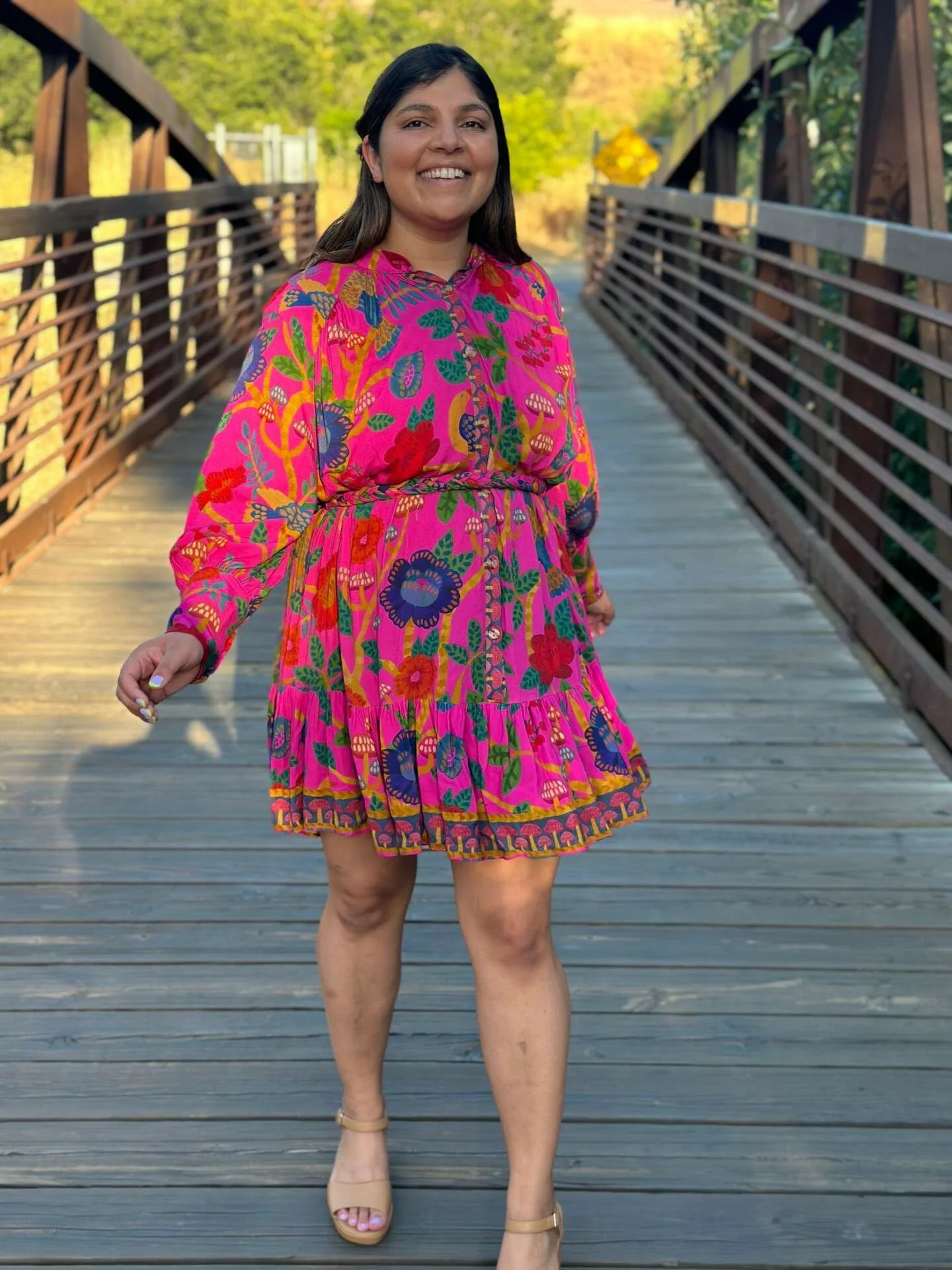 Person wearing a vibrant floral dress standing on a wooden bridge, smiling, with greenery in the background.