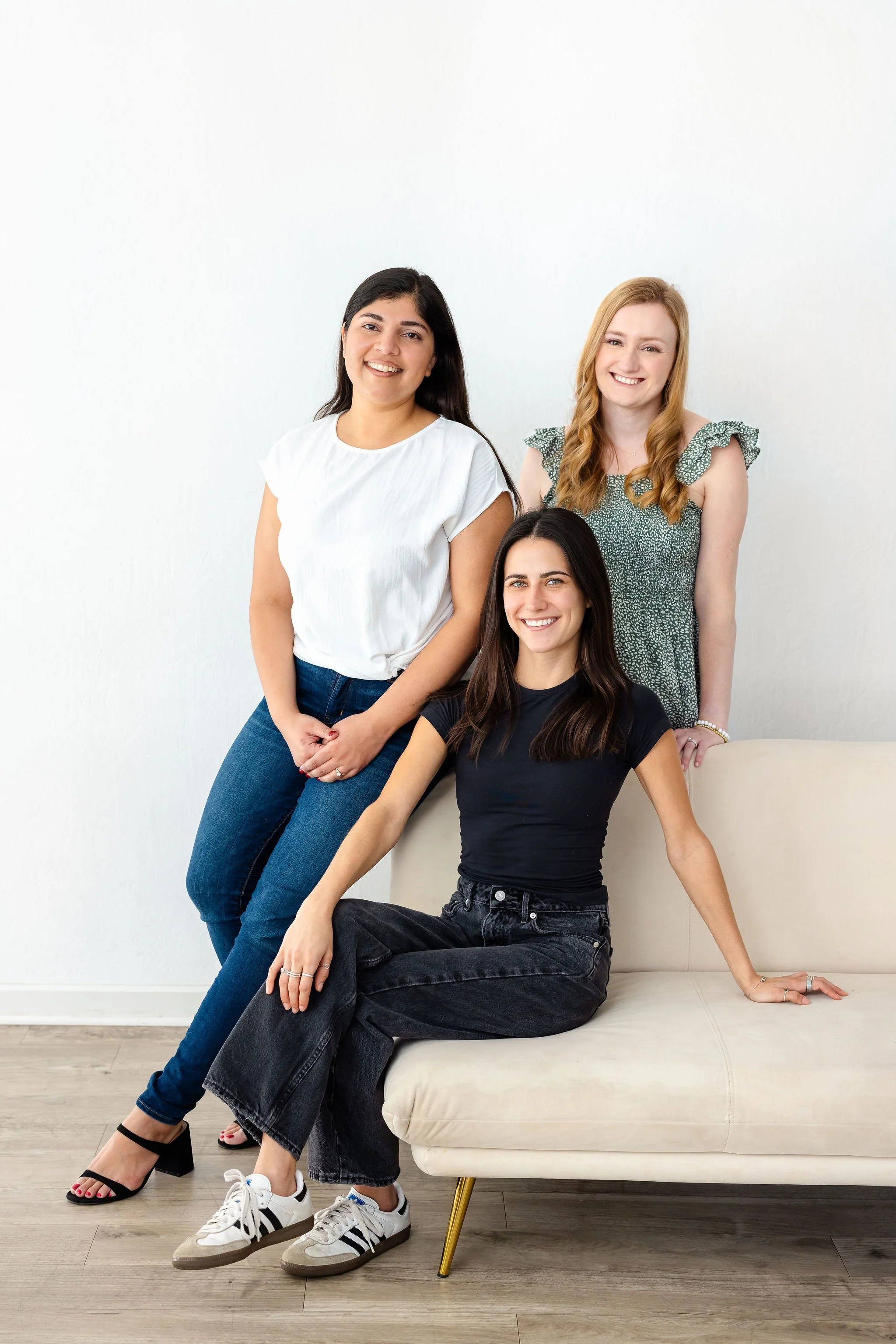 Three women posing together, two standing and one sitting on a white sofa, in casual attire against a plain white background.