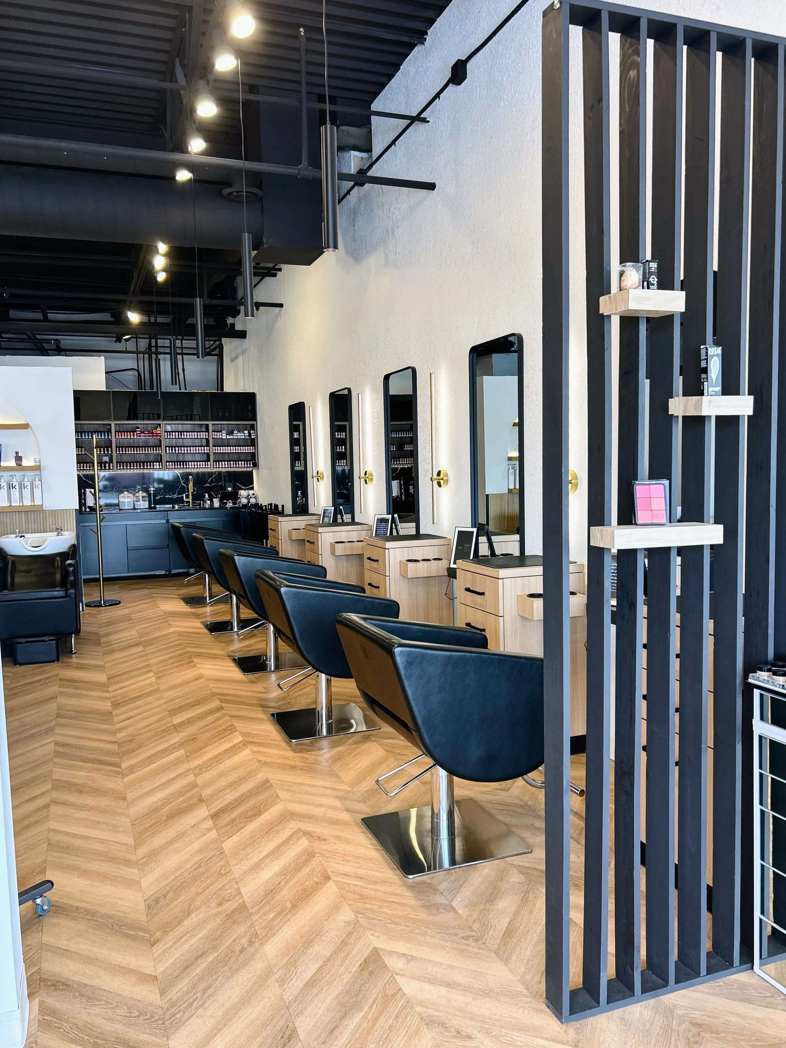 Interior of a modern hair salon with black chairs, wooden station counters, and mirrors, with shelves on the side wall displaying small products.