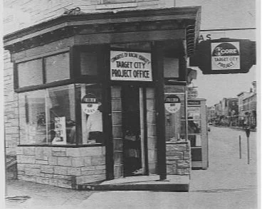 A small corner building with signs for Target City Project Office and other businesses, featuring large windows and a brick and wood exterior, located on a busy street.
