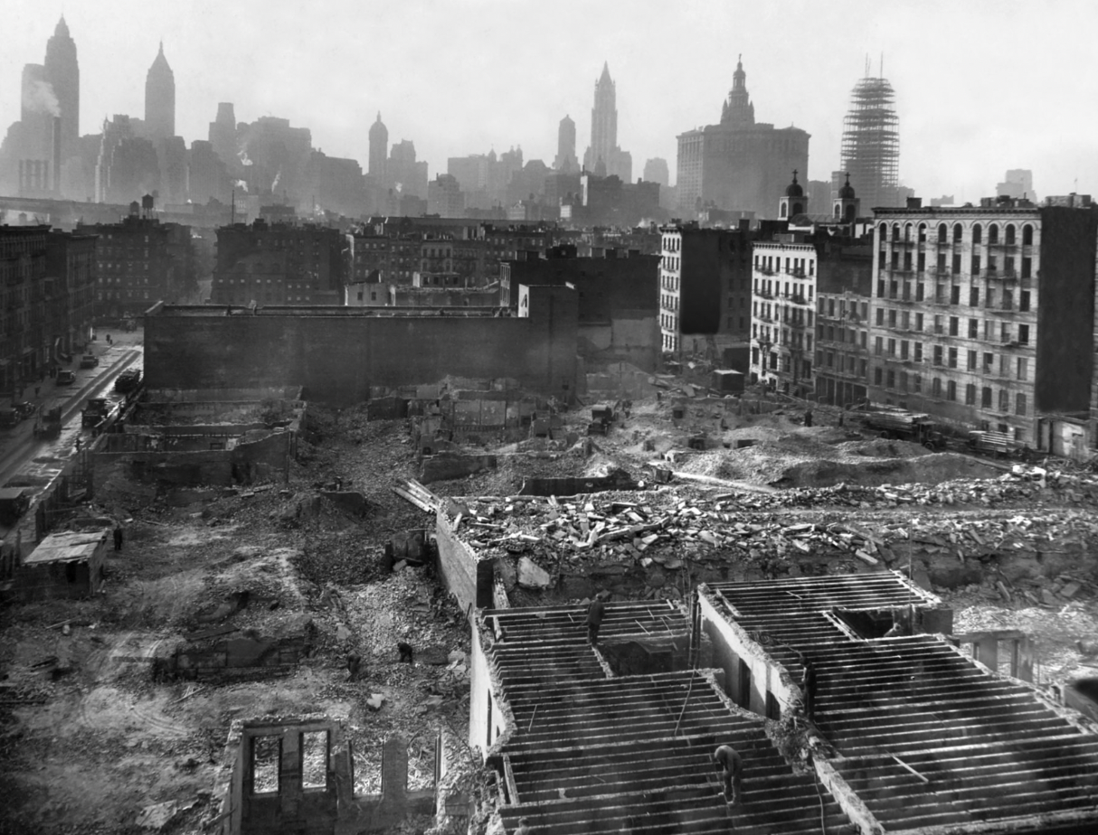 A black-and-white photo of a cityscape with numerous buildings and skyscrapers in the background, and an area in the foreground under construction or demolition with debris and visible building frameworks.