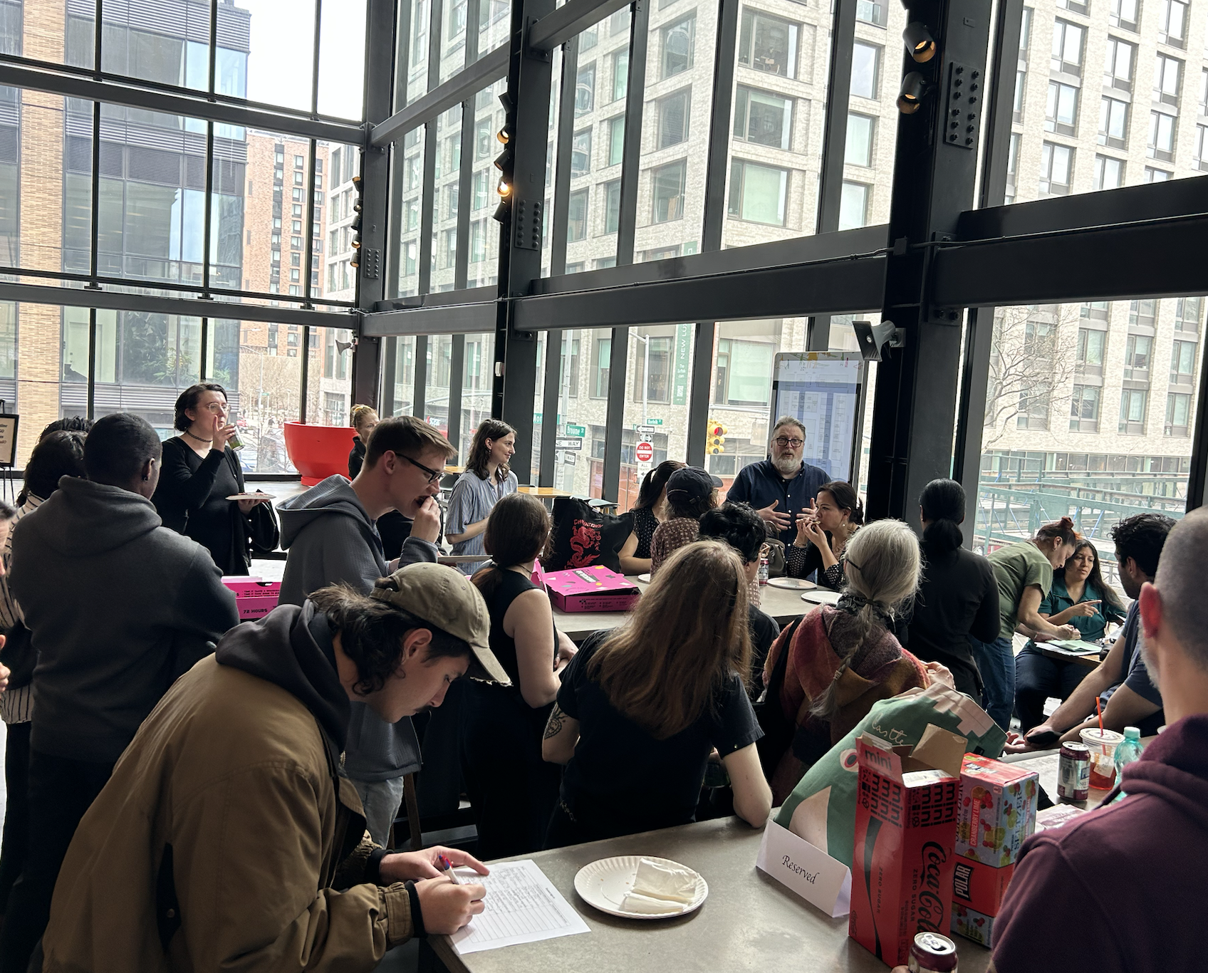 A group of people gathered around tables in a brightly lit, modern indoor space with large glass windows and a view of city buildings outside. Some individuals are engaged in conversation, while others are eating or reading. There are various food and drink items on the tables.