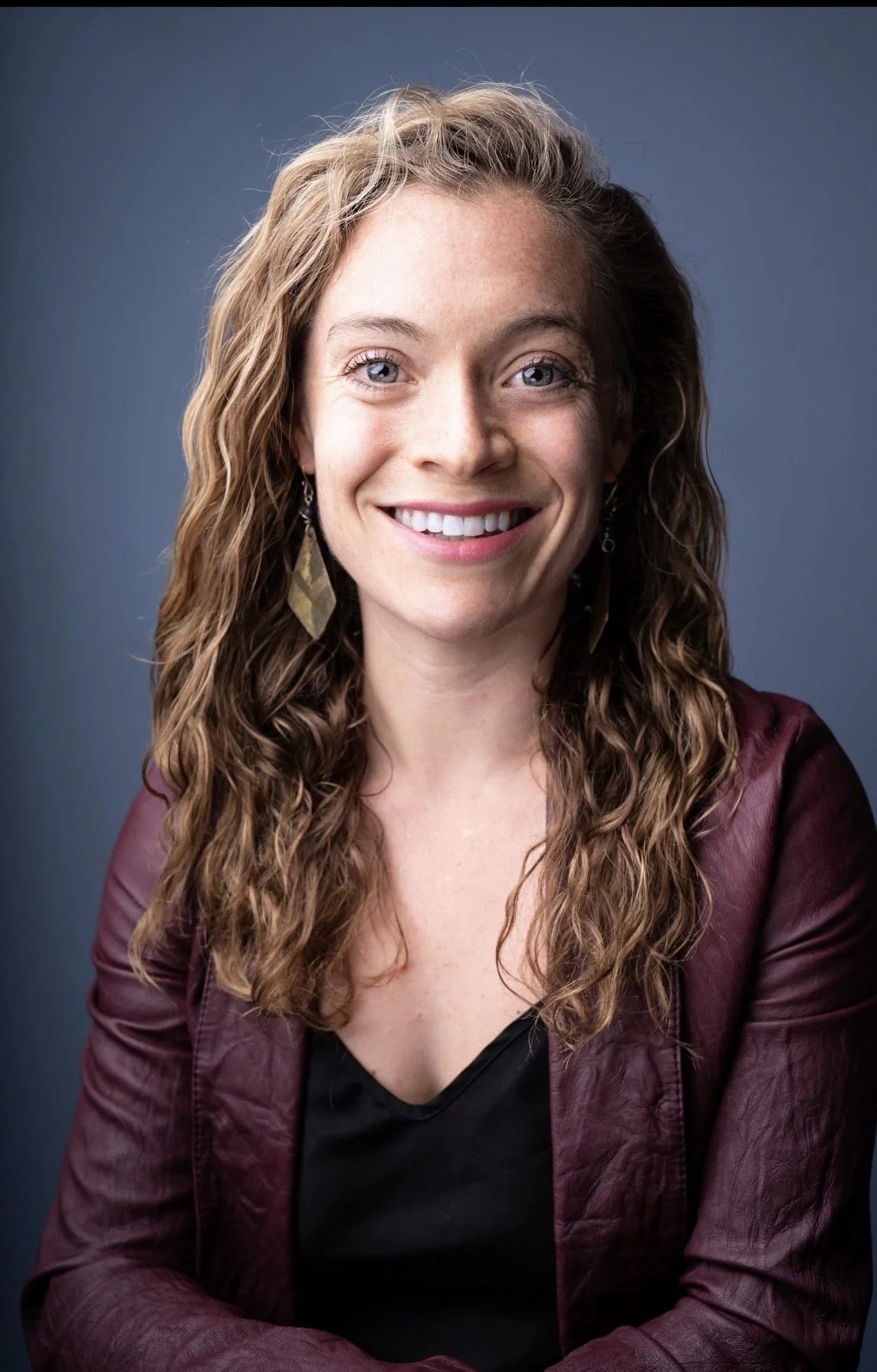 A woman with long, curly brown hair wearing earrings, a black top, and a maroon jacket, smiling against a dark blue background.