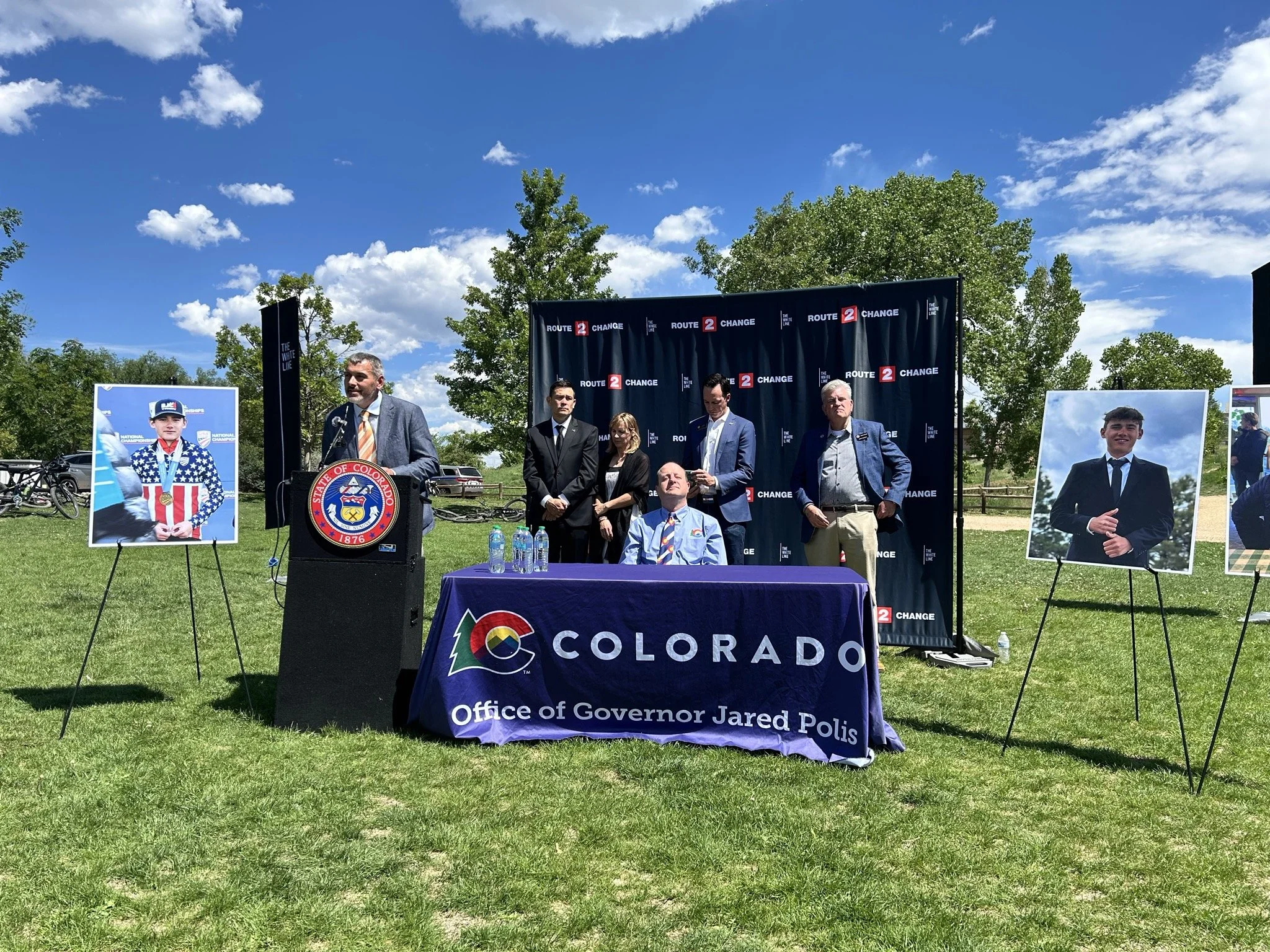 A political outdoor event with a group of people standing in front of a banner that reads 'Route 2 Change.' There are large portraits of two young men on easels on either side of the group, and a table covered with a Colorado Office of Governor Jared Polis banner. The setting is a sunny day with a blue sky and scattered clouds, surrounded by green trees.
