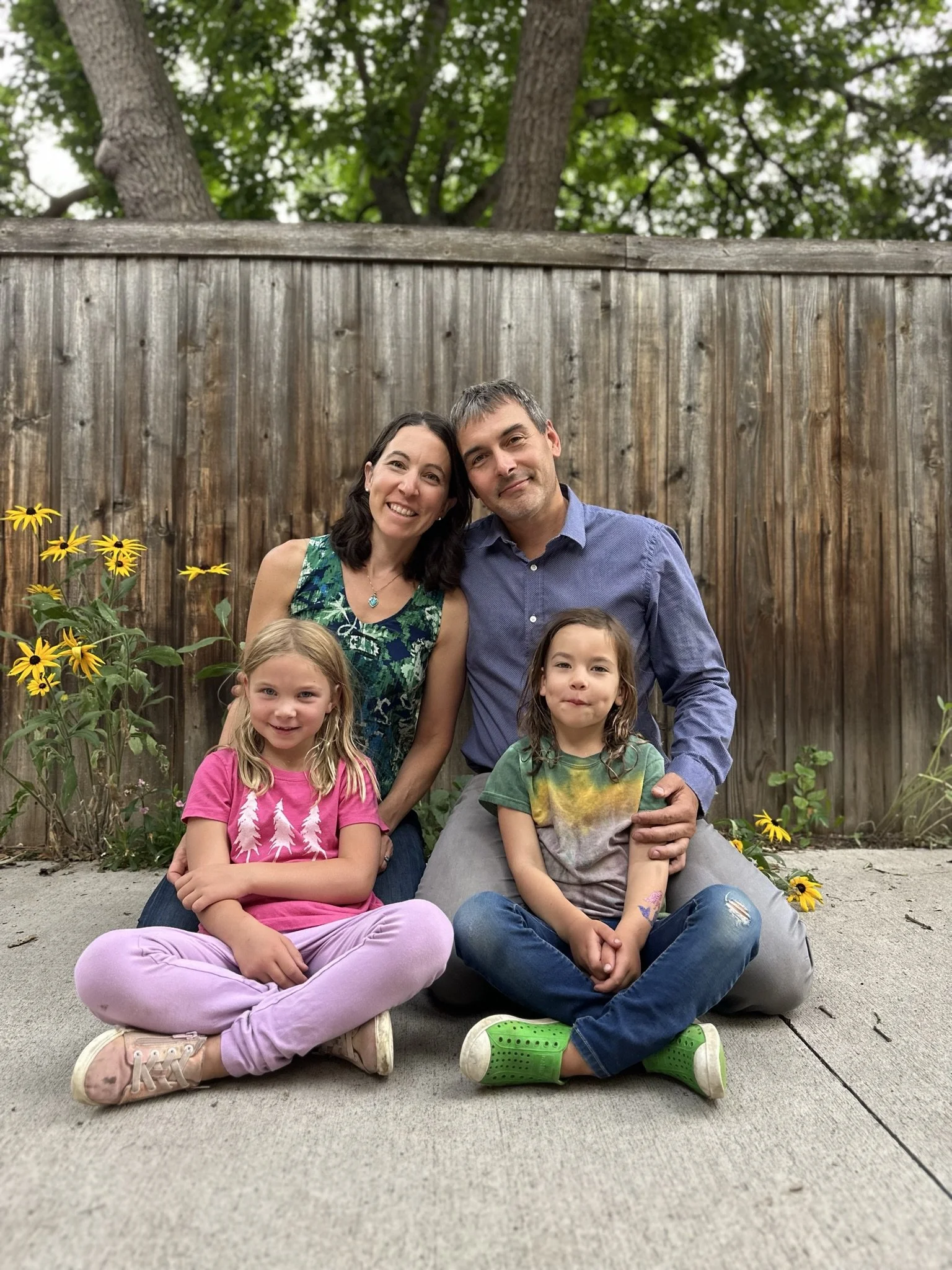 Family photo of a woman, man, and two children sitting on a concrete porch in front of a wooden fence with trees and yellow flowers in the background.