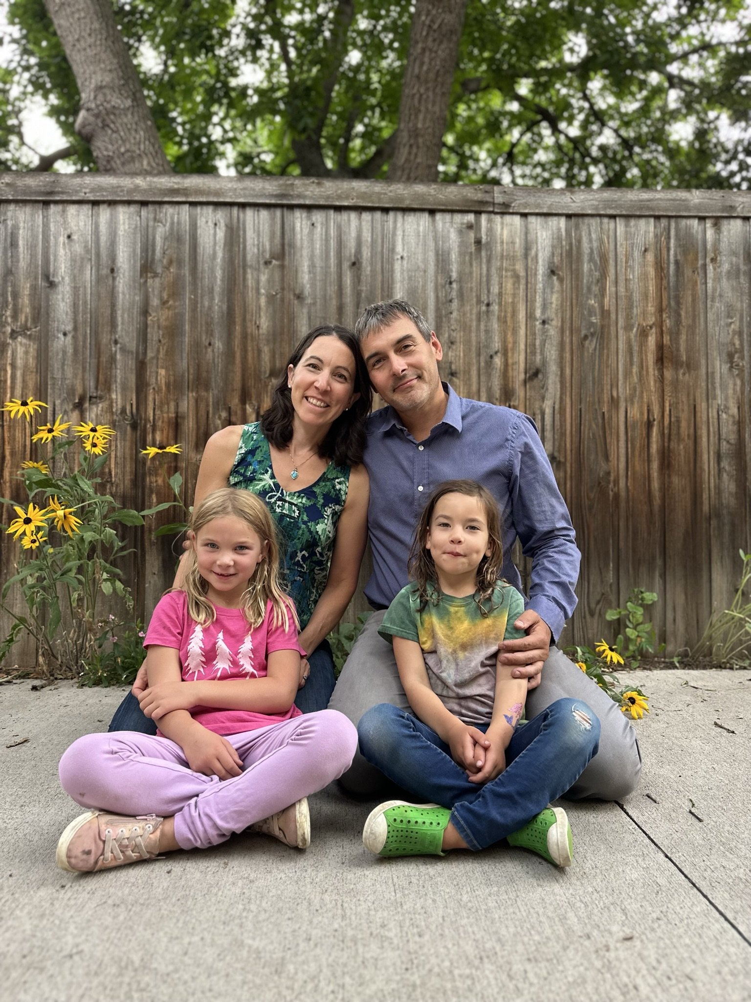 Family photo of a woman, man, and two children sitting on a concrete porch in front of a wooden fence with trees and yellow flowers in the background.