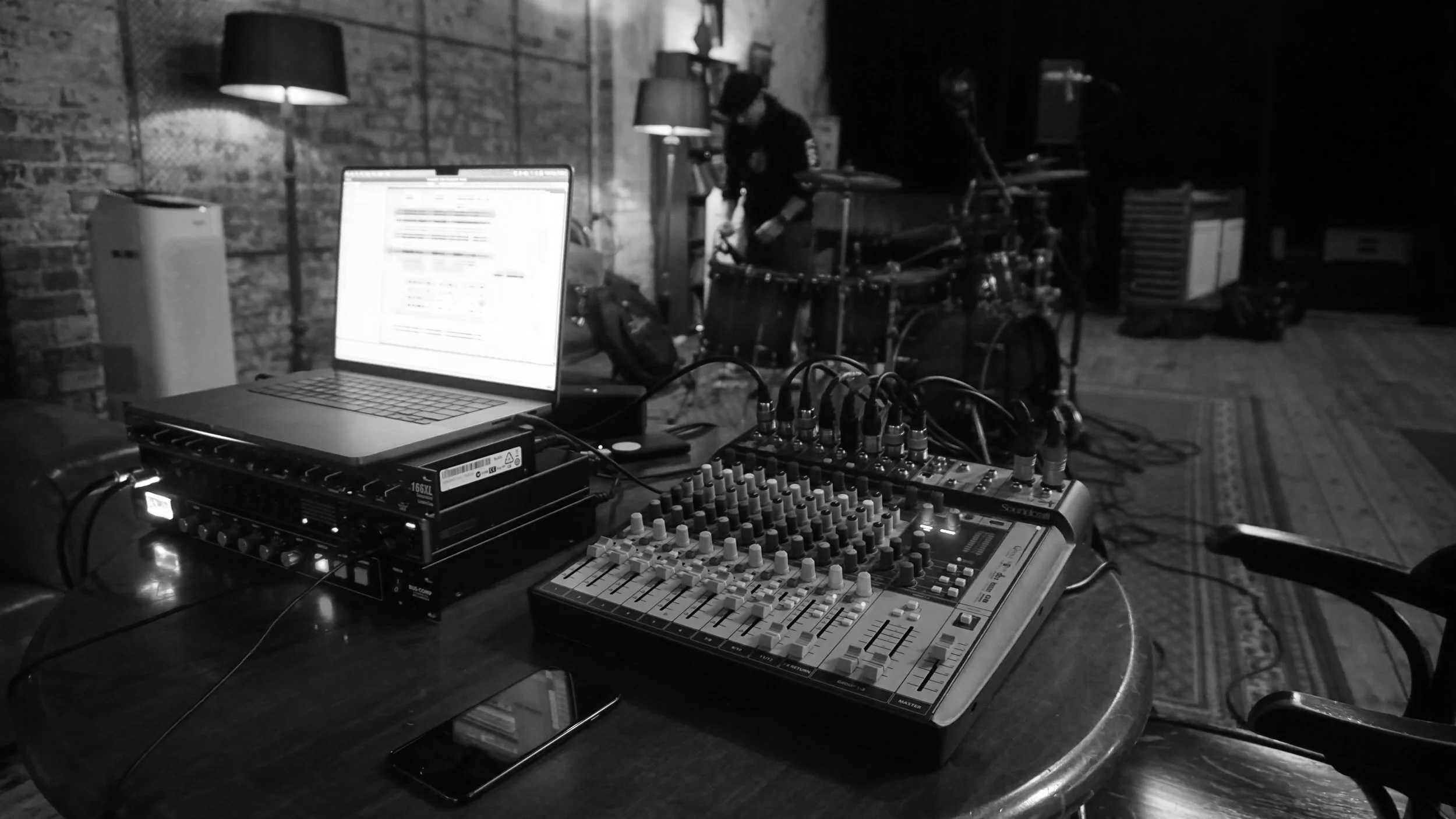 A sound mixer and laptop on a table, with a person setting up music equipment in a dimly lit room with brick walls and floor lamps.