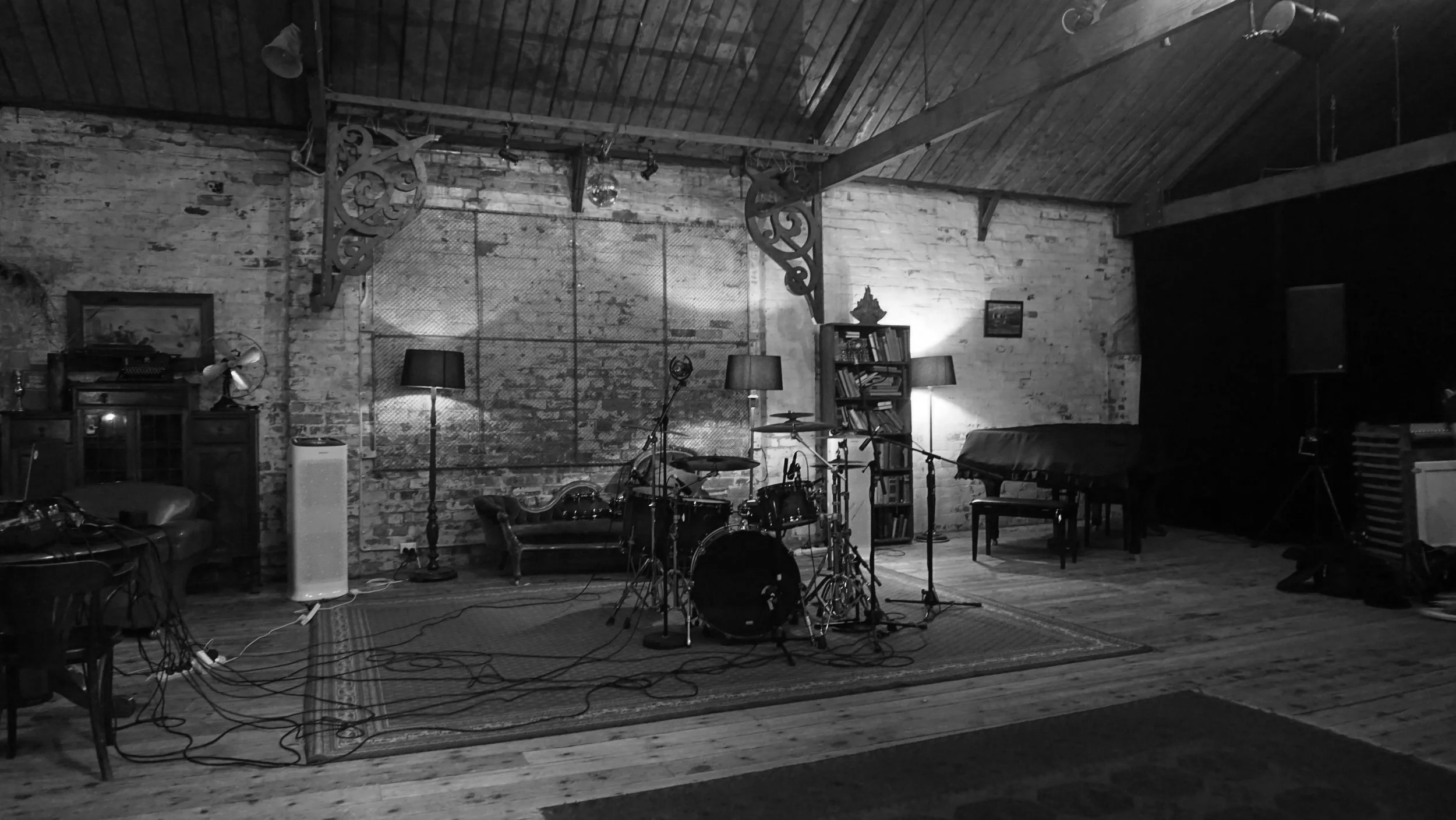 An indoor music studio with exposed brick walls, a drum set, microphones, and various musical equipment arranged on a wooden floor with a patterned rug.