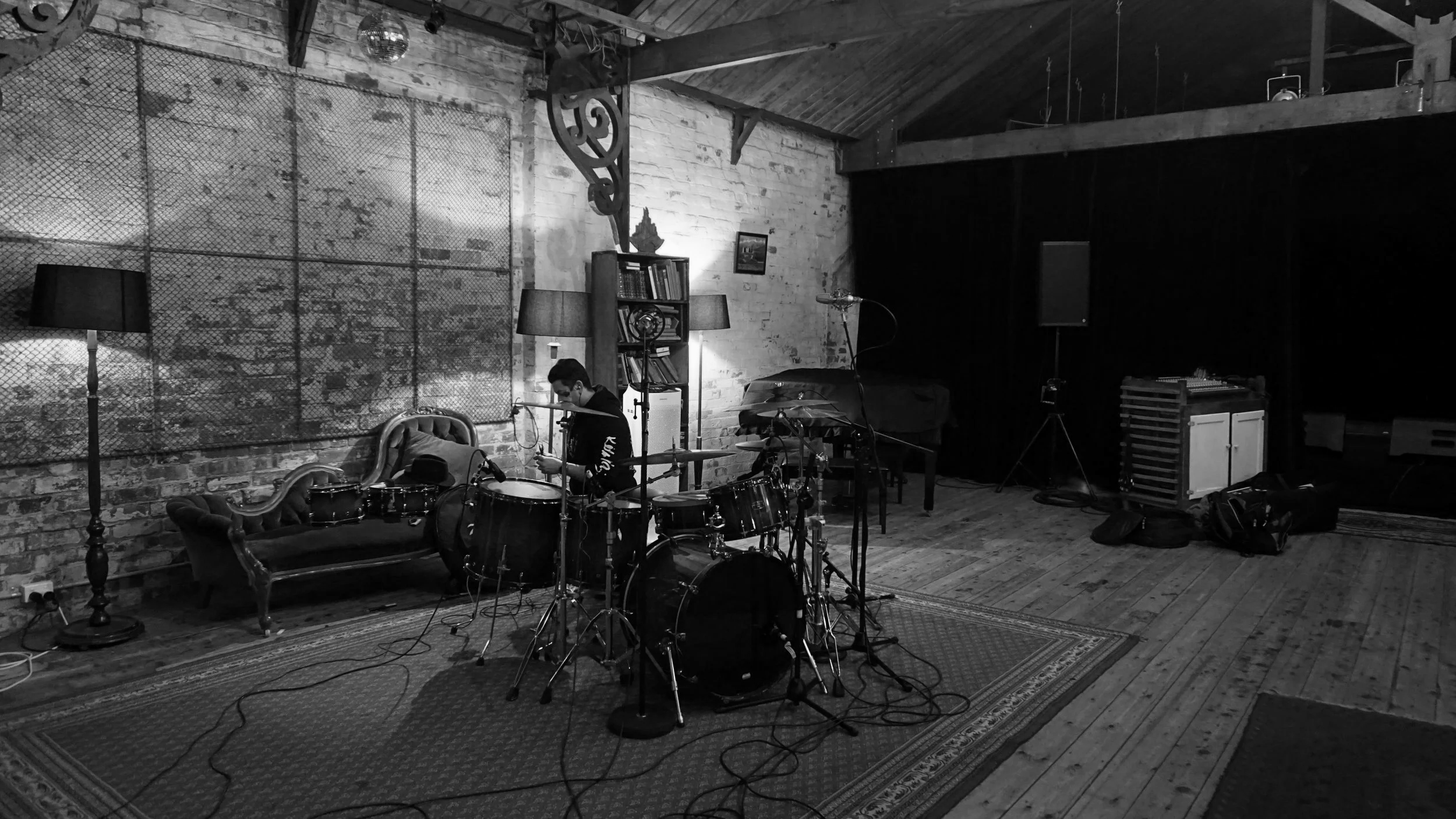 A musician setting up drums on a stage in an industrial-style venue with brick walls, wooden beams, furniture, and stage equipment in black and white.