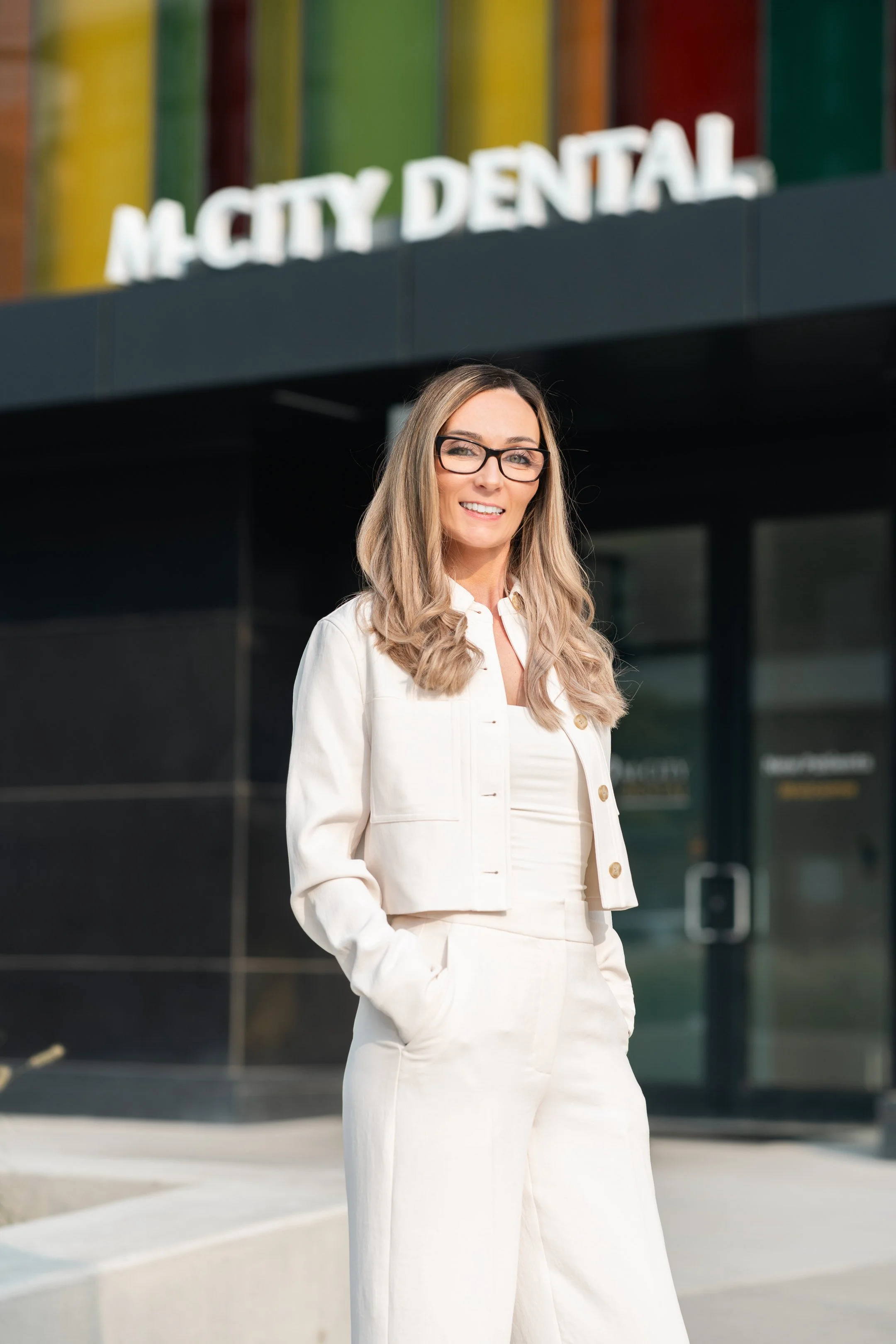 A woman (Sandra Harrower) with long blonde hair and glasses standing outside a building with a sign that reads 'M CITY DENTAL'.