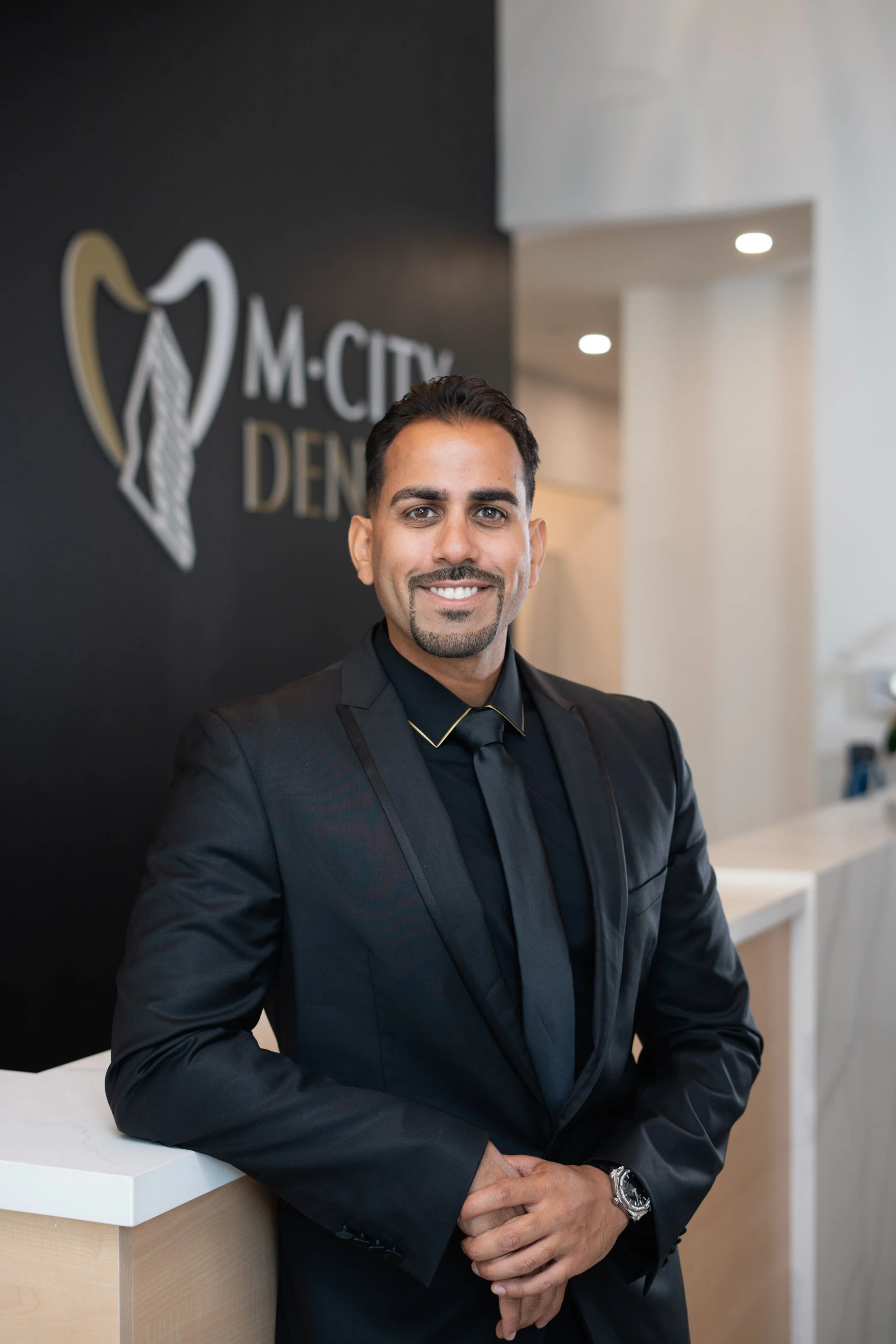 A dentist (Dr. Mahmoud Manzoor) in a black suit and tie smiling at the camera, standing in front of a reception desk with a sign that reads 'M-CITY DENTAL' in a professional office setting.