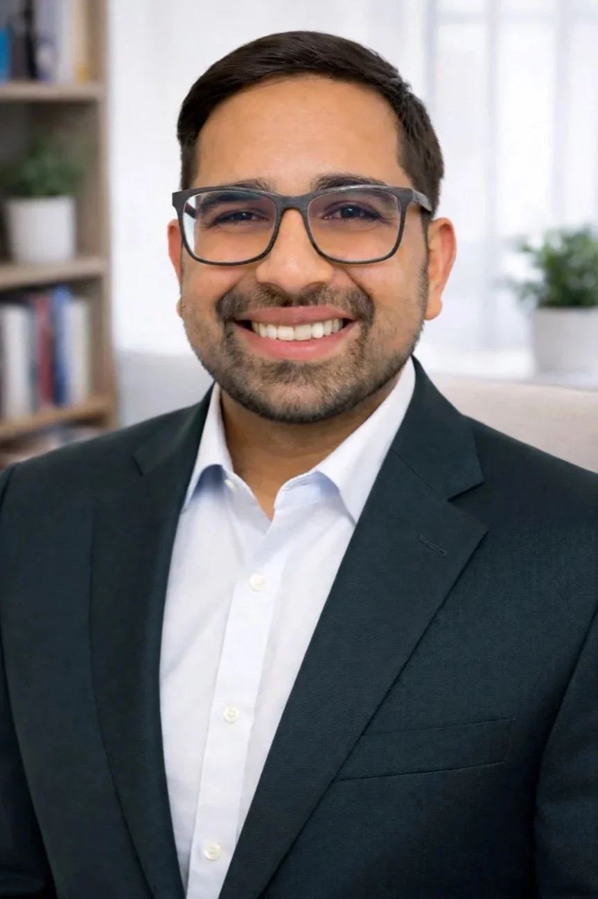 A smiling man in a black suit, white shirt, and glasses, sitting in a room with books and plants in the background.