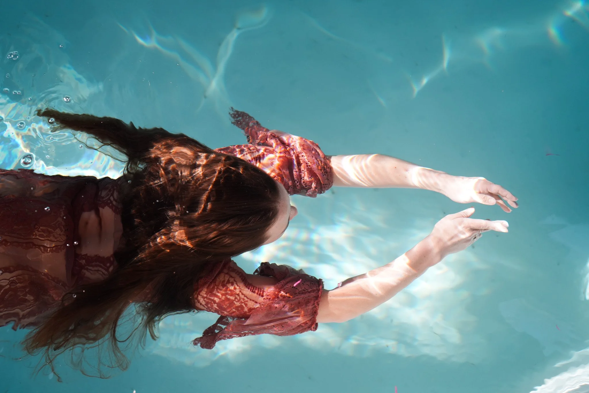 A woman with long brown hair in a lace red dress swimming underwater in a pool with her arms stretched forward.