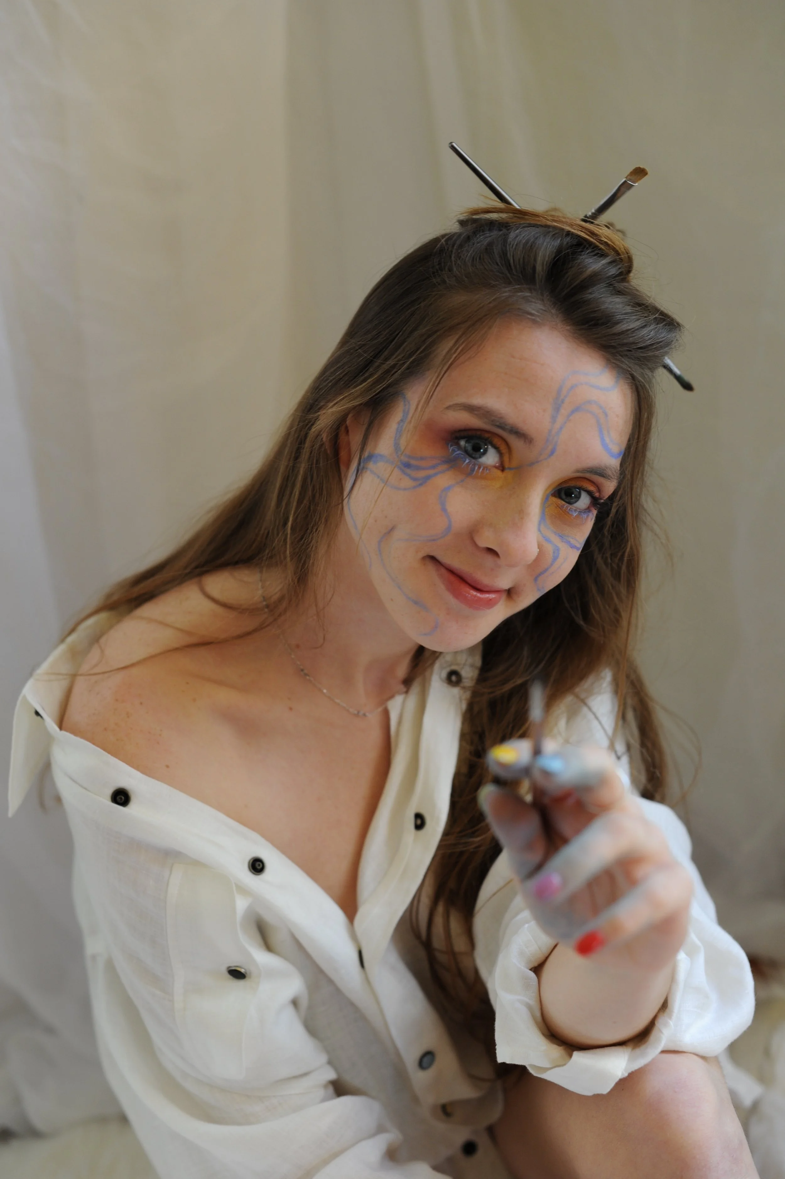 Monet Poe has colorful face paint with blue swirl lines. She is holding a paintbrush and smiling, with blue paint on her fingers, wearing a white shirt with rolled-up sleeves and colorful buttons.