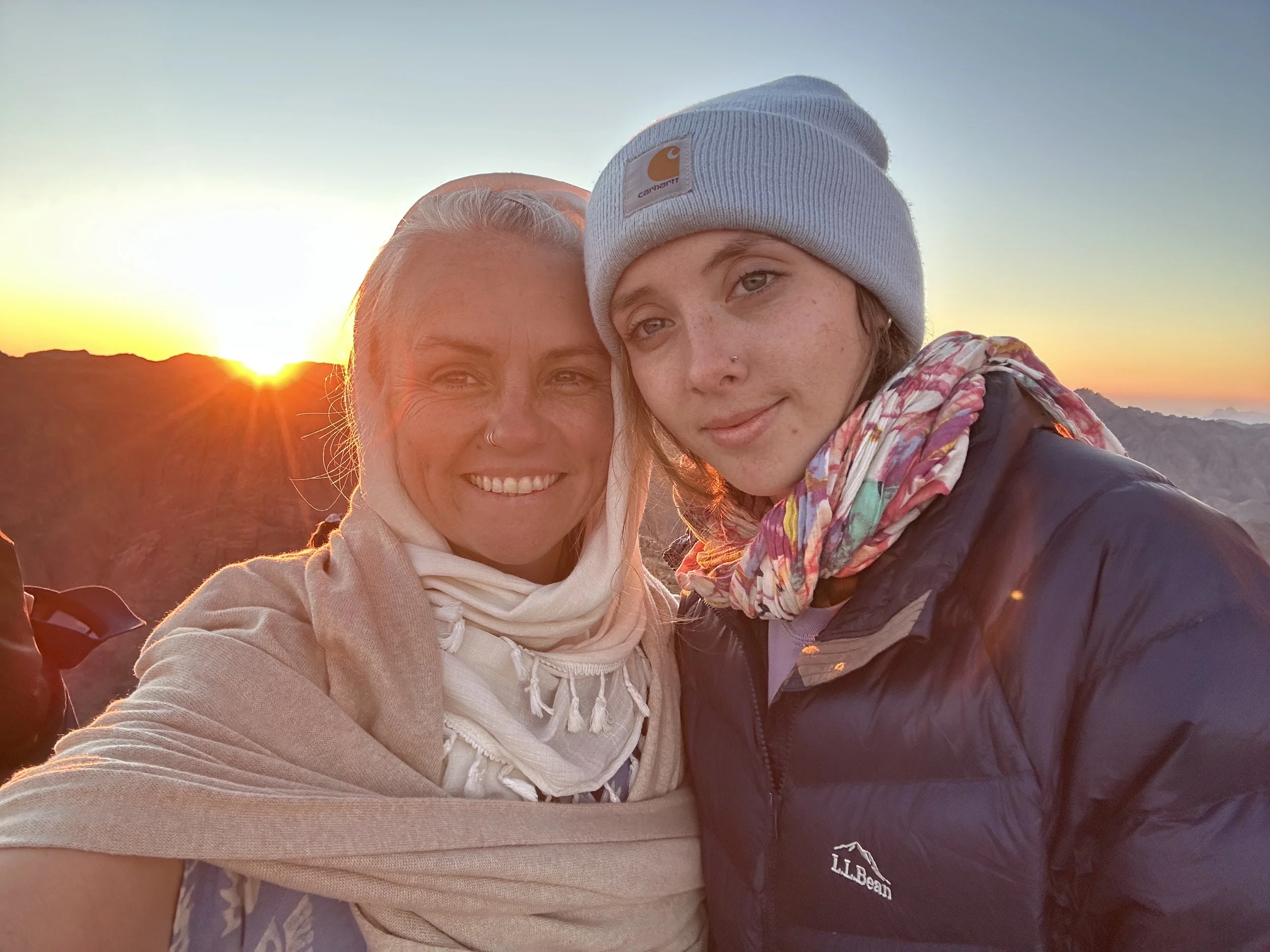 Sarah Poe and Monet Poe, mother daughter taking a selfie outdoors at sunset with mountains in the background, one with gray hair and a beige scarf, and the other with a beanie, colorful scarf, and a navy jacket.