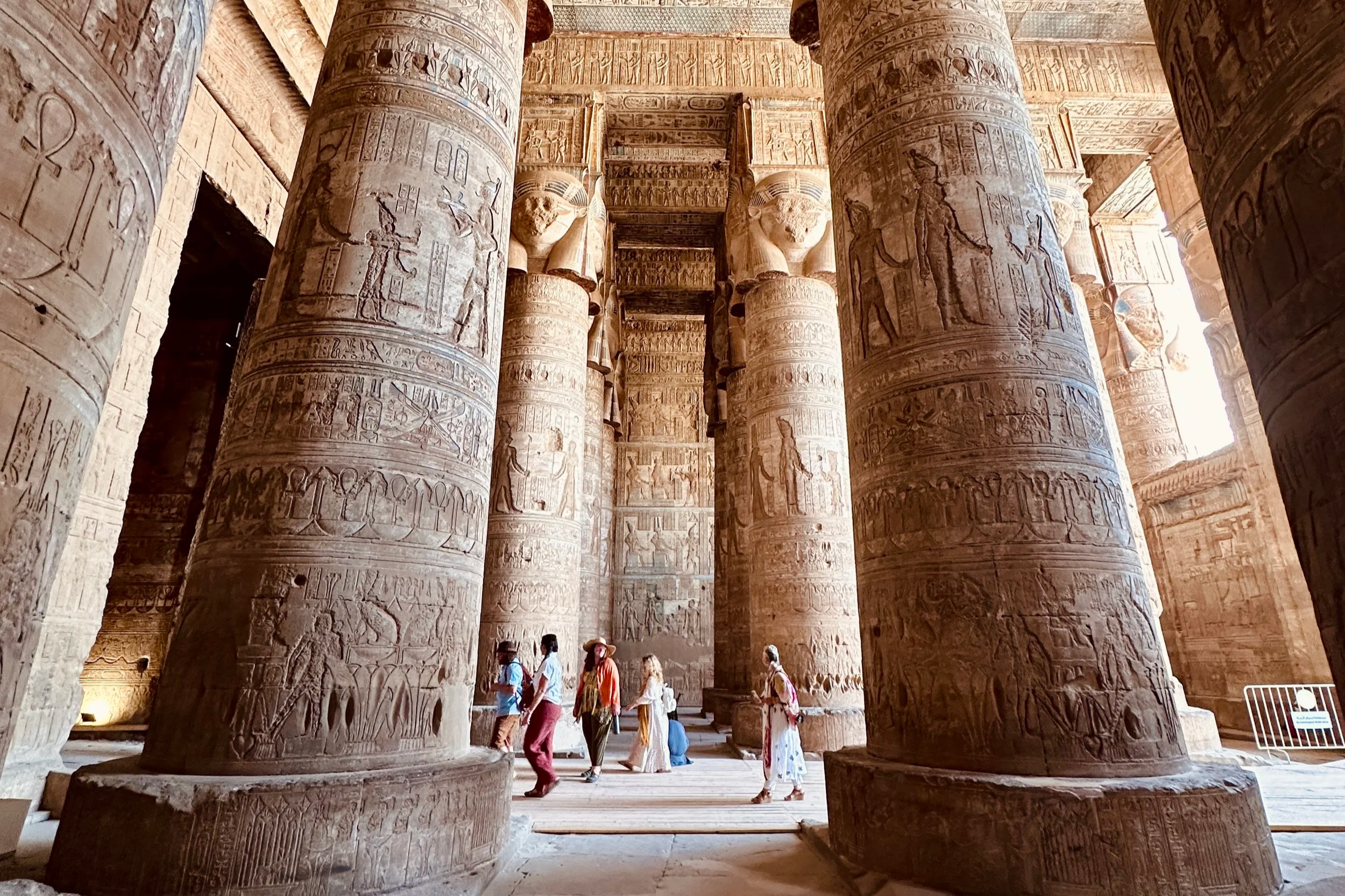 Ancient Egyptian temple interior with large hieroglyphic-covered columns and a group of tourists exploring.
