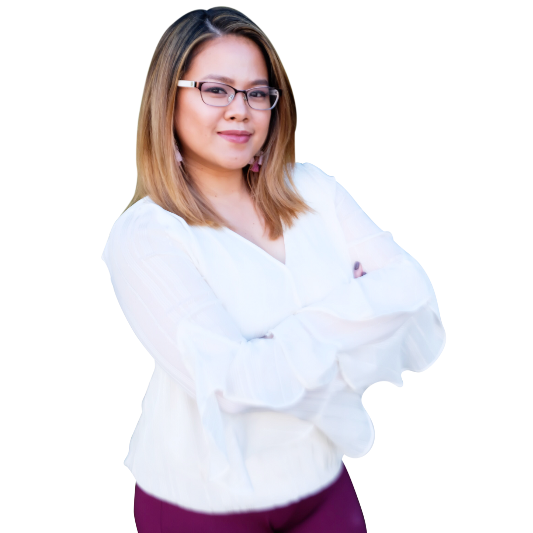A woman with shoulder-length light brown hair, wearing glasses, a white blouse with long sleeves, and purple pants, stands with her arms crossed and looks confidently at the camera.
