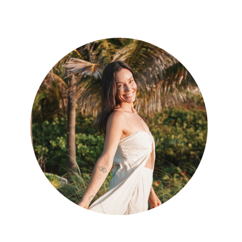 A smiling woman in a white dress standing outdoors with palm trees in the background.