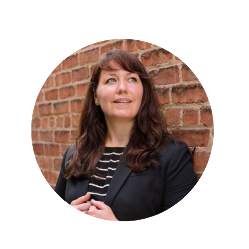 A woman with brown hair and a black blazer smiling while standing against a brick wall.