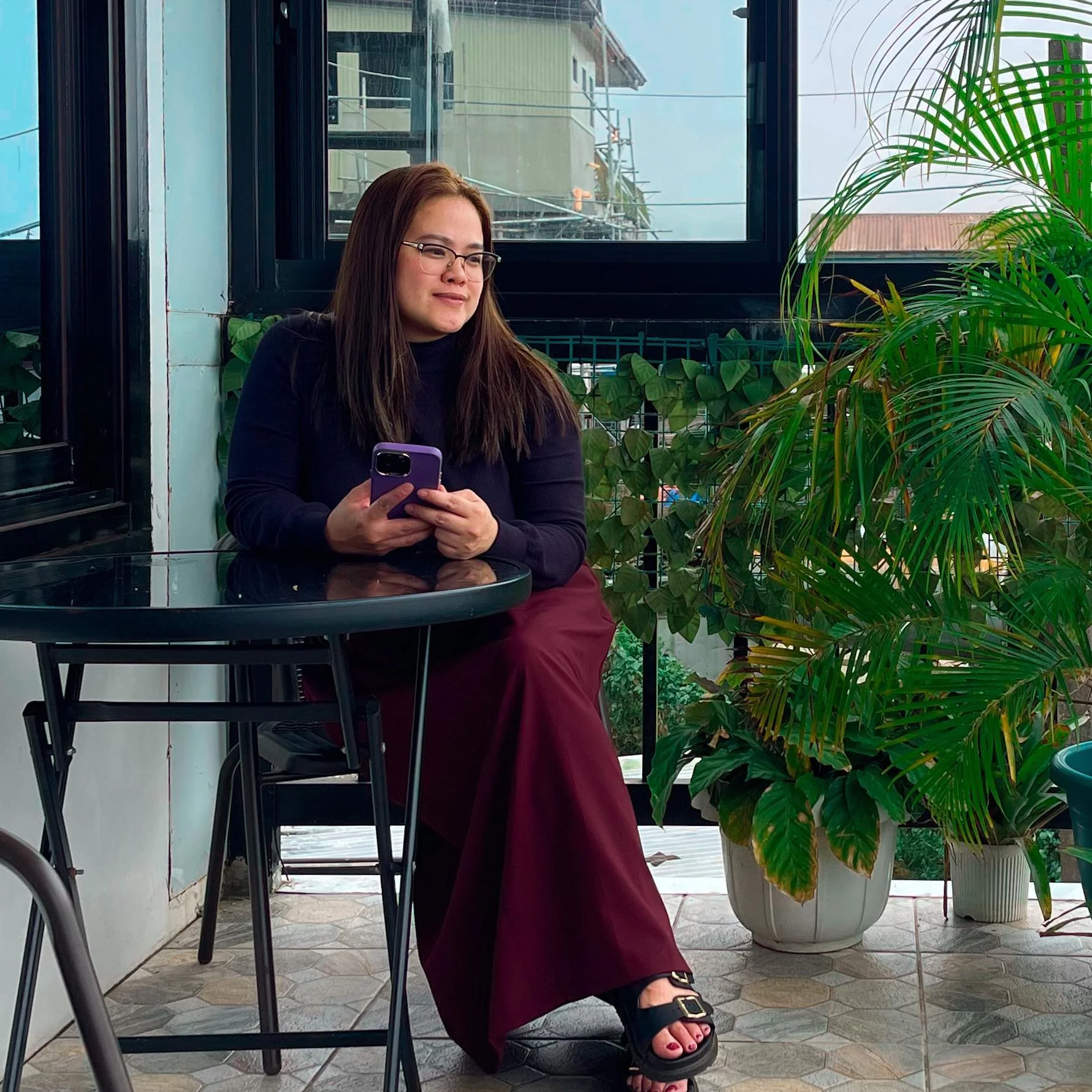 A woman sitting at a black round table on an outdoor patio, holding a smartphone, with large green potted plants and a window in the background.