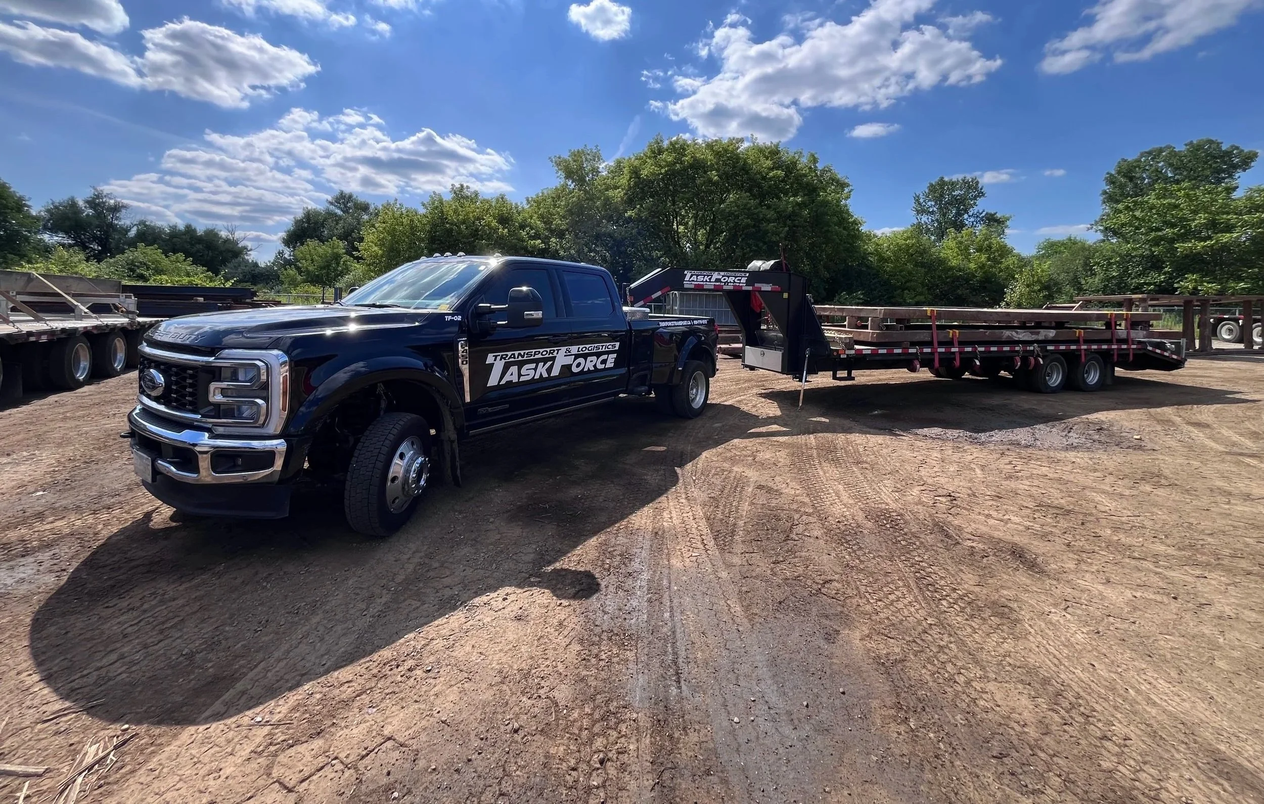 Black transport truck with logo 'Task Force' on the door, connected to a long flatbed trailer, parked on dirt ground, with a partly cloudy sky and green trees in the background.