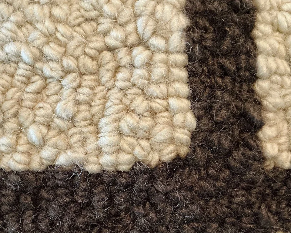 Detail of hand-hooked wool loops showing tonal variation between oat ground and umber cross.