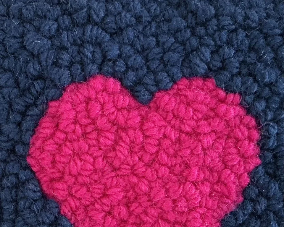 Close-up of hand-hooked wool loops showing a bright pink heart against a navy ground.