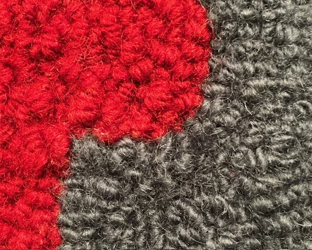 Close-up of hand-hooked wool loops in red and charcoal, showing dense texture and fiber variation.