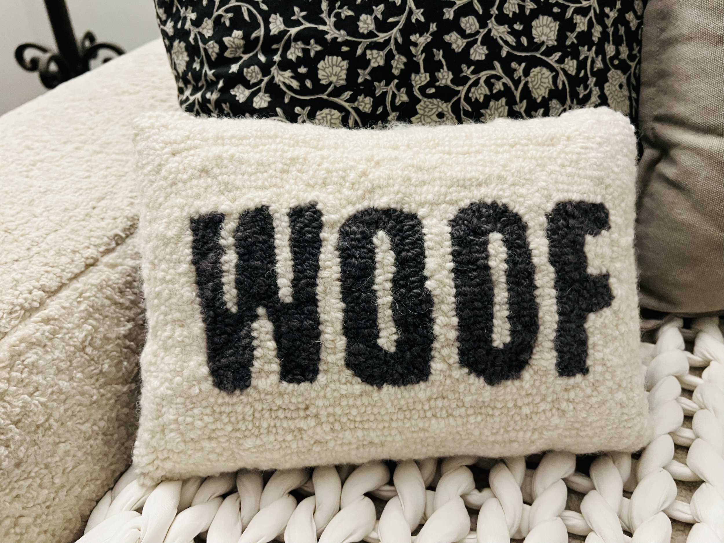 A small beige textured pillow with the word 'WOW' in large black embroidery, placed on a woven white surface, with additional pillows in patterned and plain fabric in the background.