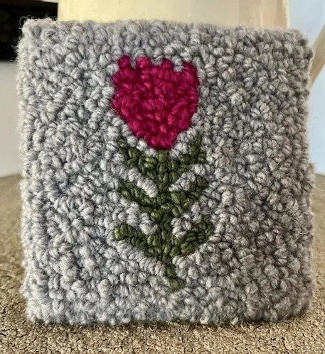 A gray fuzzy square pillow with an embroidered pink heart-shaped flower with green stem and leaves in the center.