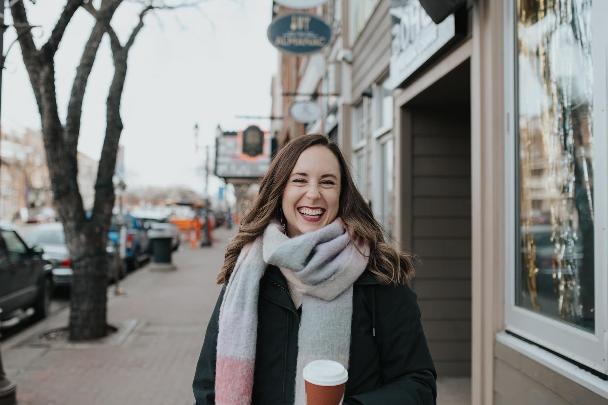 A woman smiling and holding a coffee to go on a city sidewalk, with trees and storefronts visible in the background.