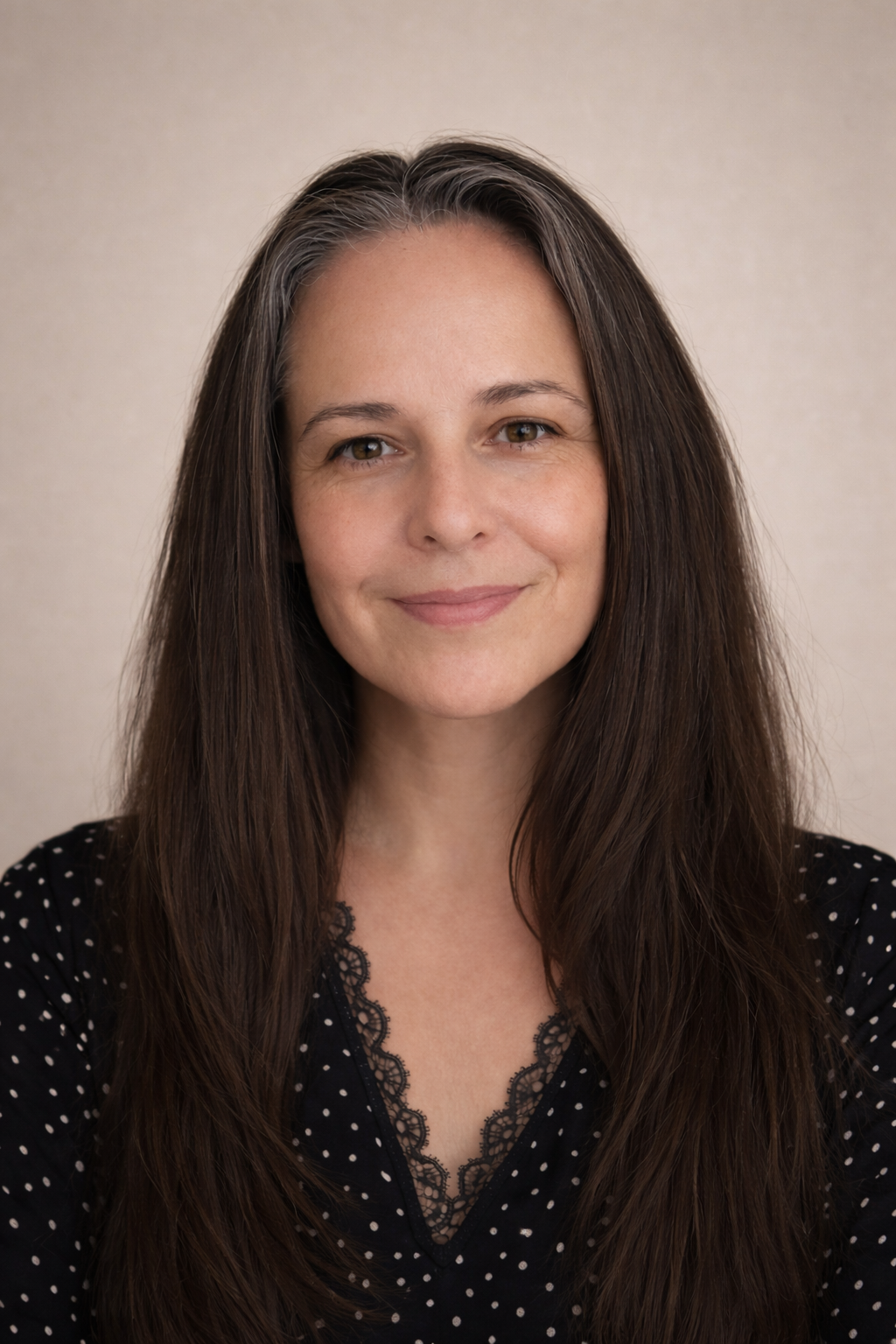 A woman with long brown hair smiling at the camera, wearing a black polka dot top with lace trim at the neckline, standing against a beige background.