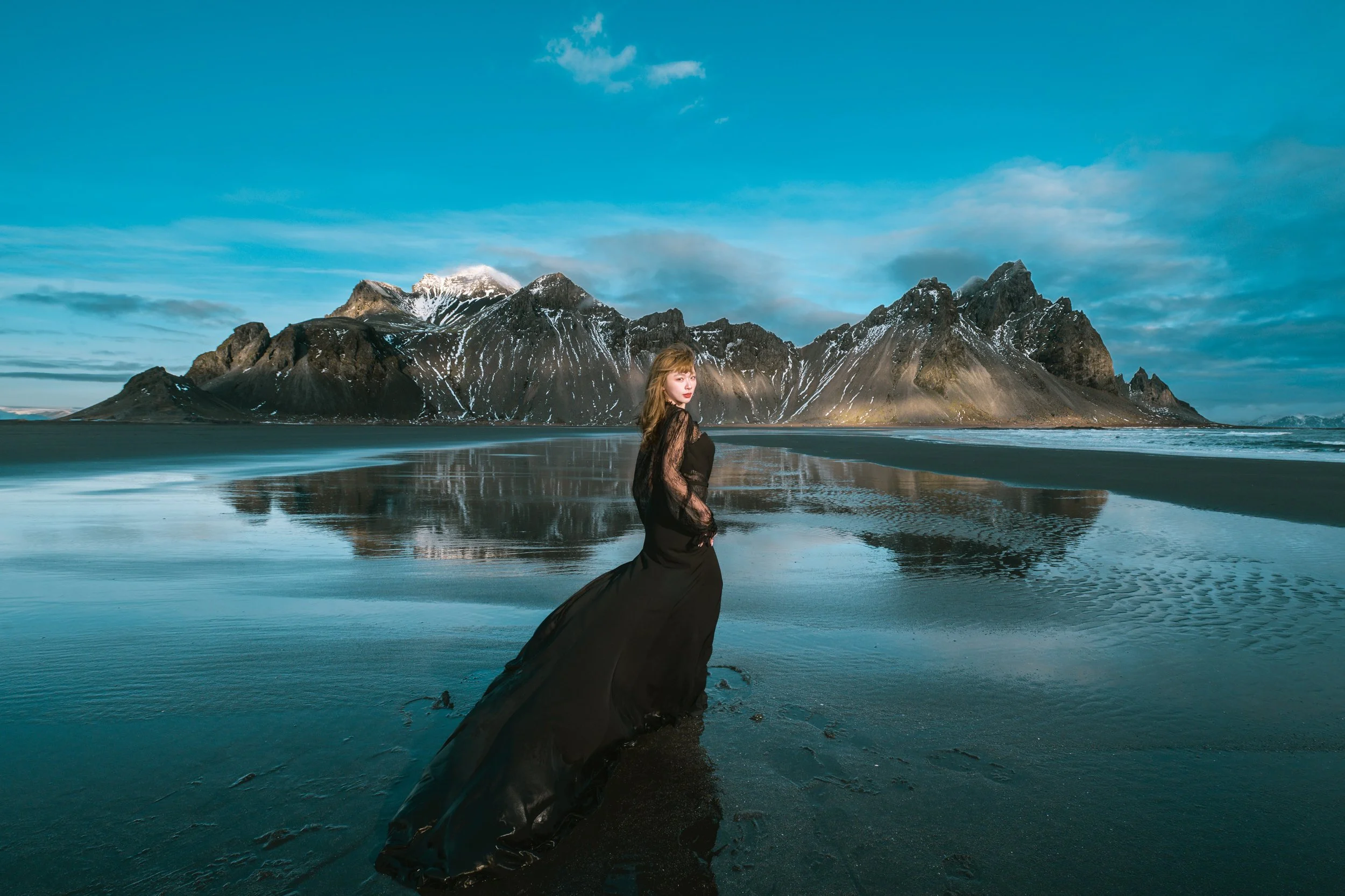 Woman in black gown standing on the wet sand with mountain reflection