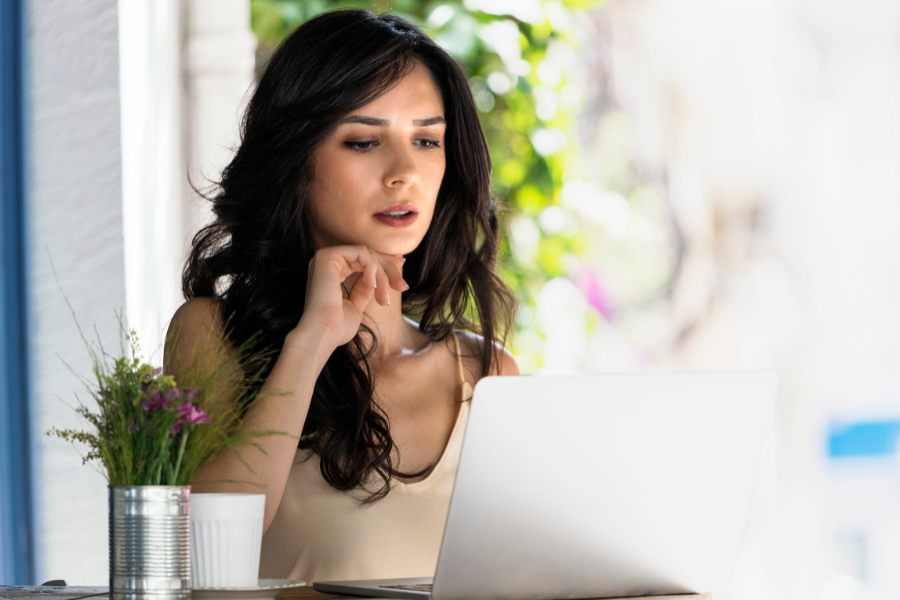Young woman with dark hair sitting at a table and looking at a laptop, with a potted plant next to her, in a bright outdoor setting.